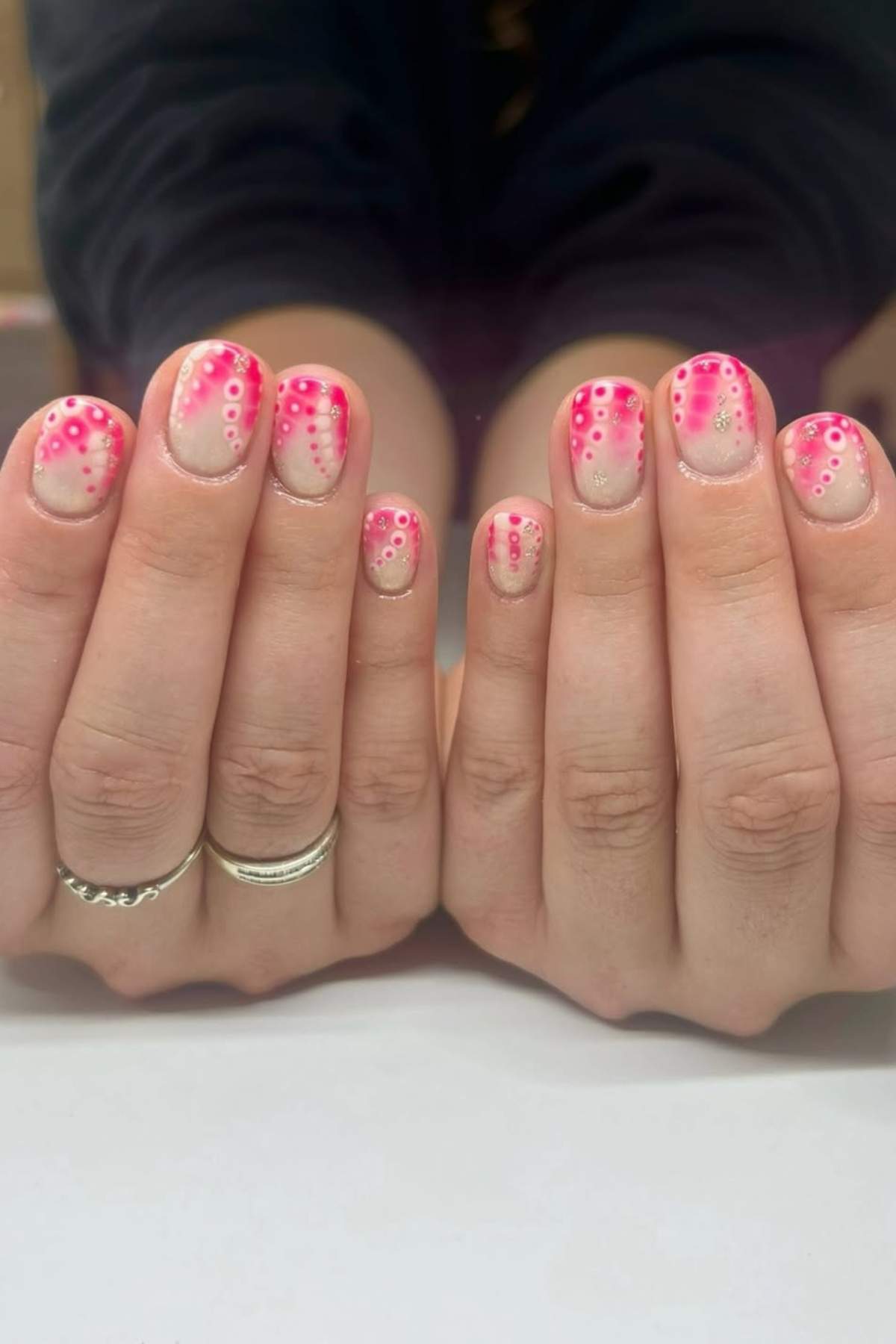 Hands with short nails painted with a light base and decorated with pink dots and rings. The person is wearing silver rings on several fingers.