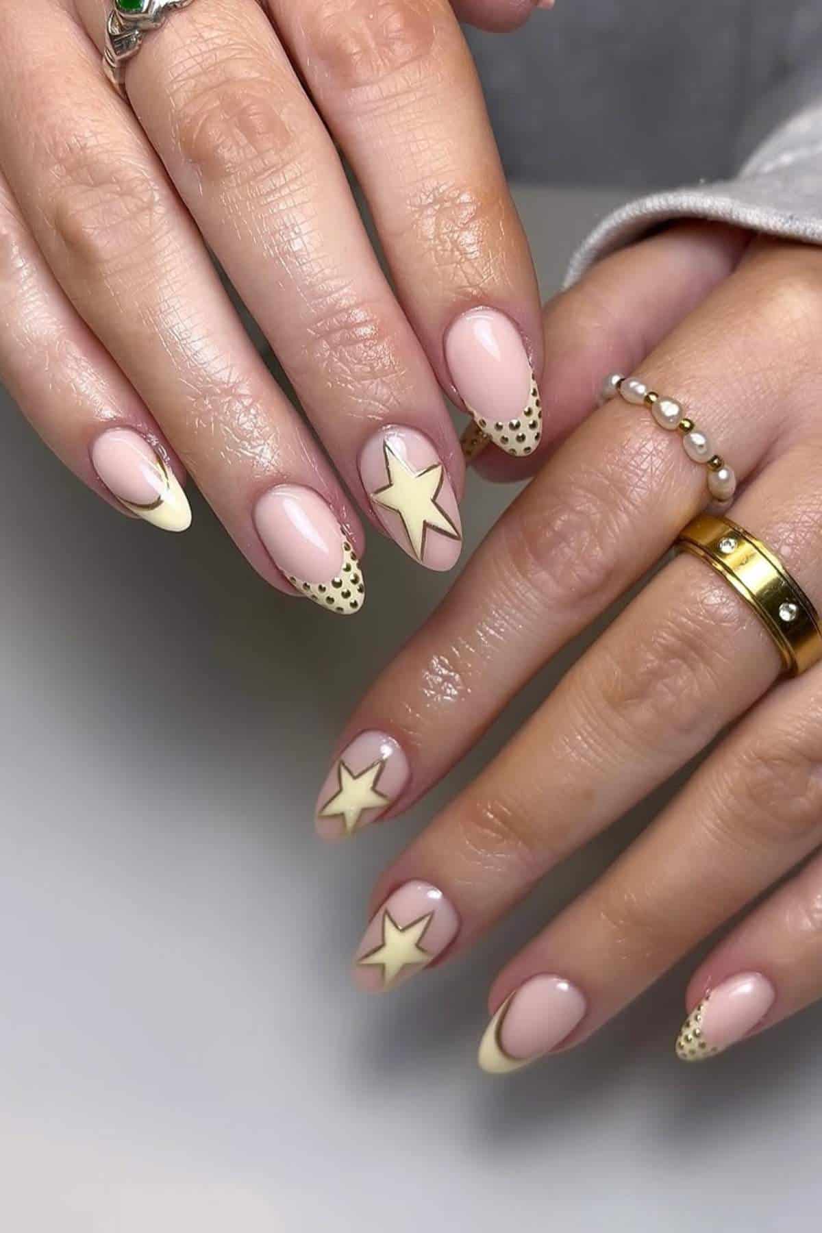Close-up of hands with long almond-shaped nails featuring a nude base, yellow star designs, cream-colored tips, and black dotted accents.