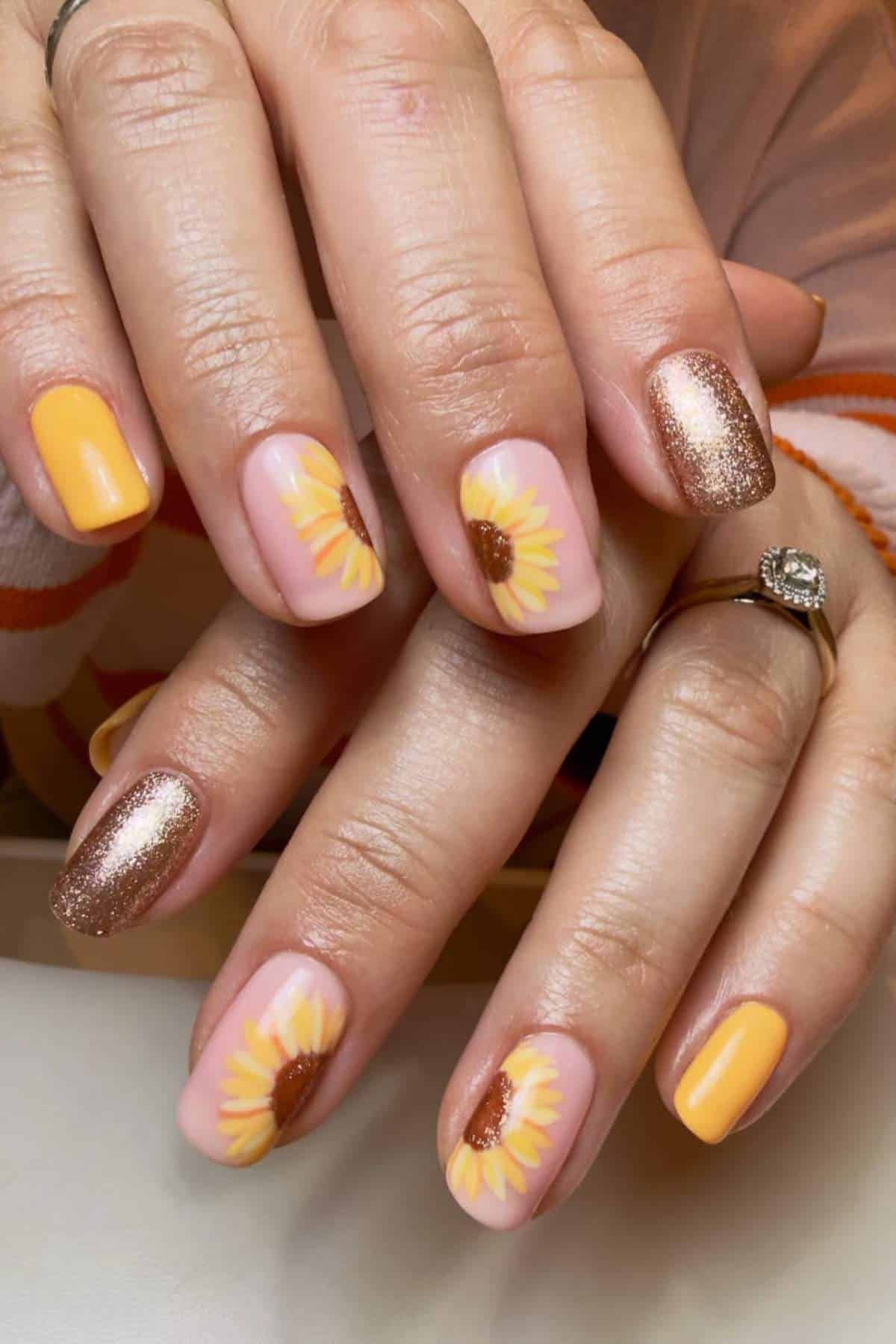 A hand with manicured nails featuring yellow sunflowers, solid yellow polish, and gold glitter accents.