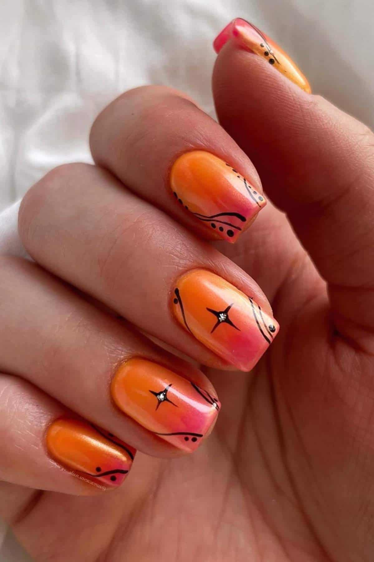 A hand with square-shaped nails painted in an orange and pink gradient design, decorated with black lines, dots, and white star accents.