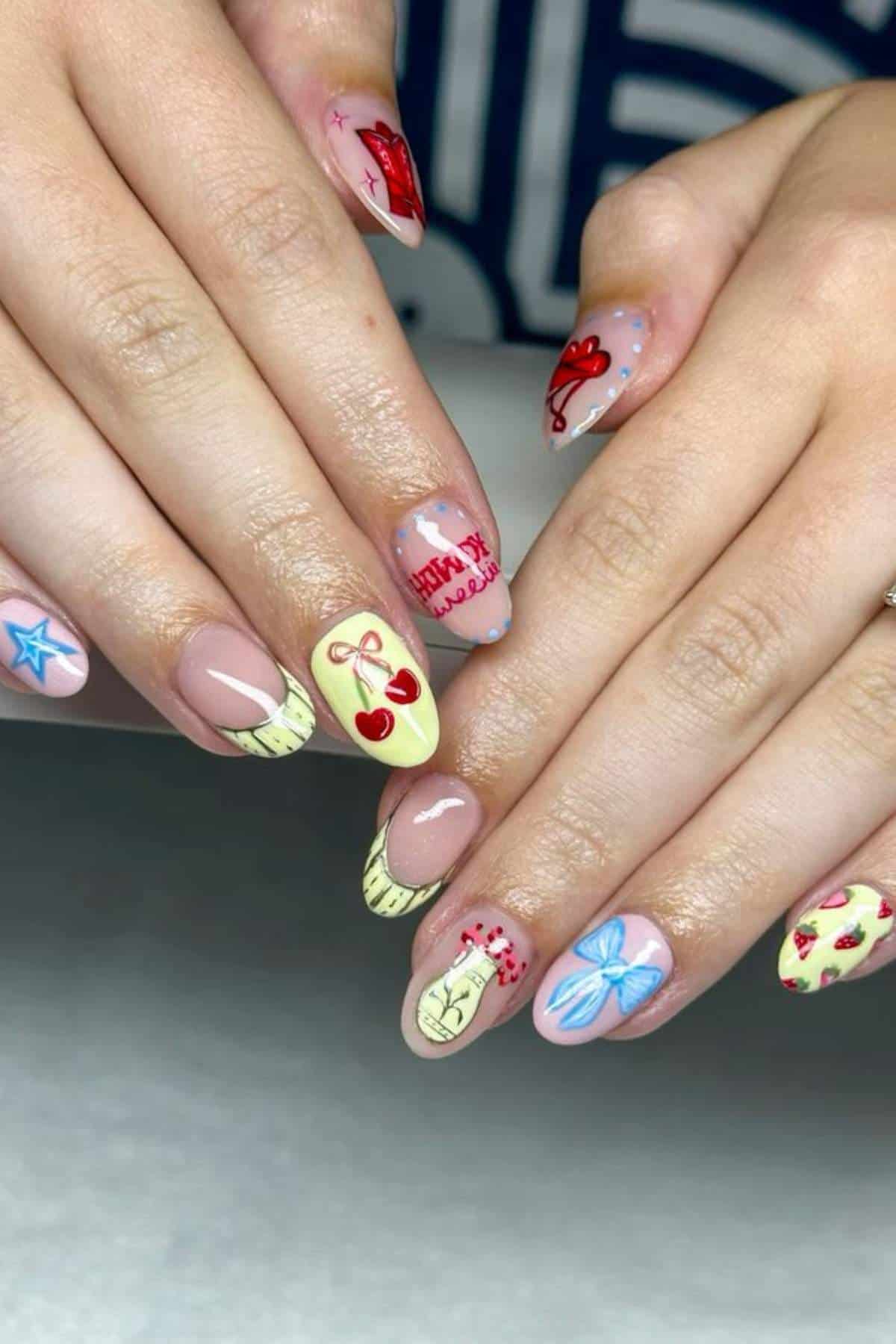 Hands with manicured nails featuring colorful designs, including cherries, stars, bows, hearts, and text on a light and nude base.