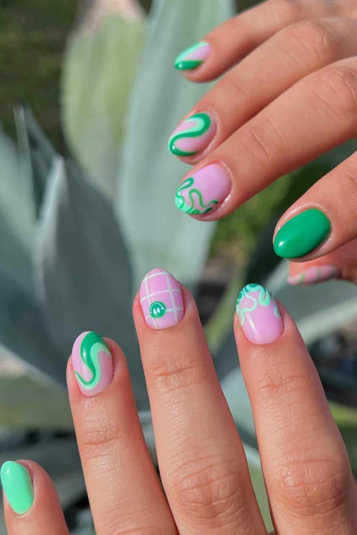 Close-up of hands with manicured nails featuring a pink and green abstract design, including swirls, plaid patterns, and solid green accents.