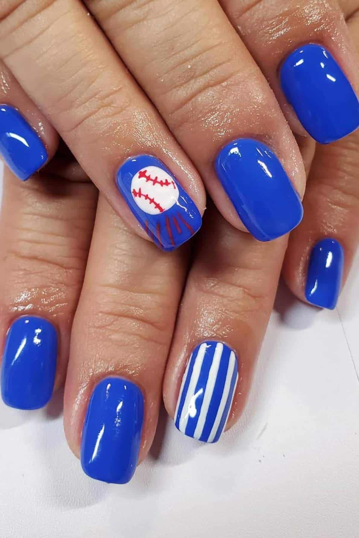 Hands with blue painted nails, one nail featuring a white baseball with red stitching, and another nail with white and blue vertical stripes.