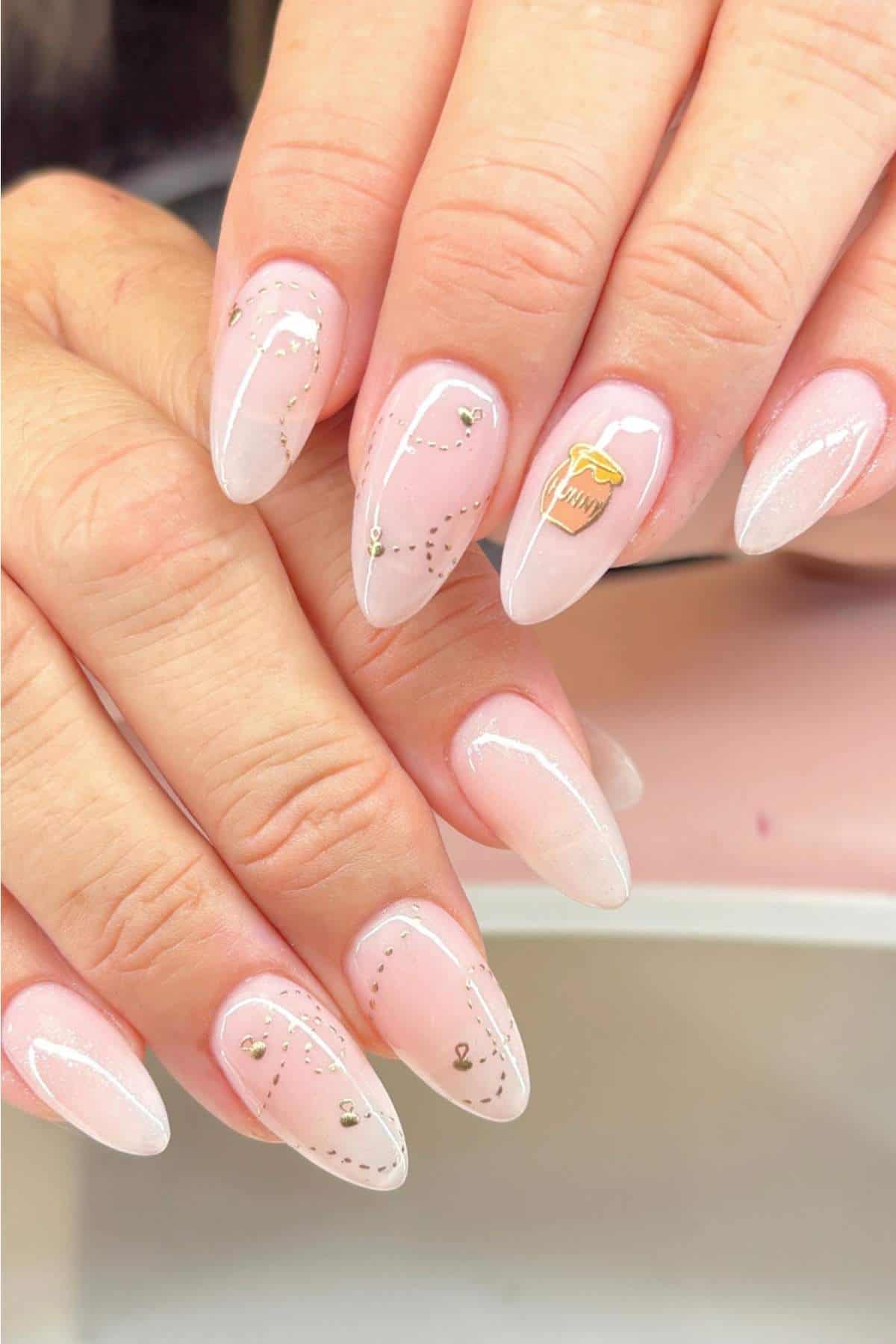 Hands with almond-shaped nails featuring a pale pink ombre manicure, delicate silver line designs, and a gold apple sticker accent on one nail.