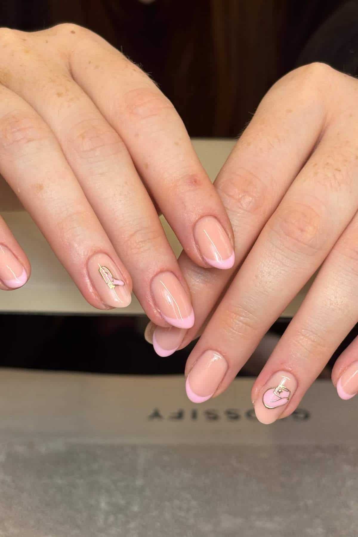 Hands with light pink French tip manicure; ring finger on each hand features a small gold ribbon design.