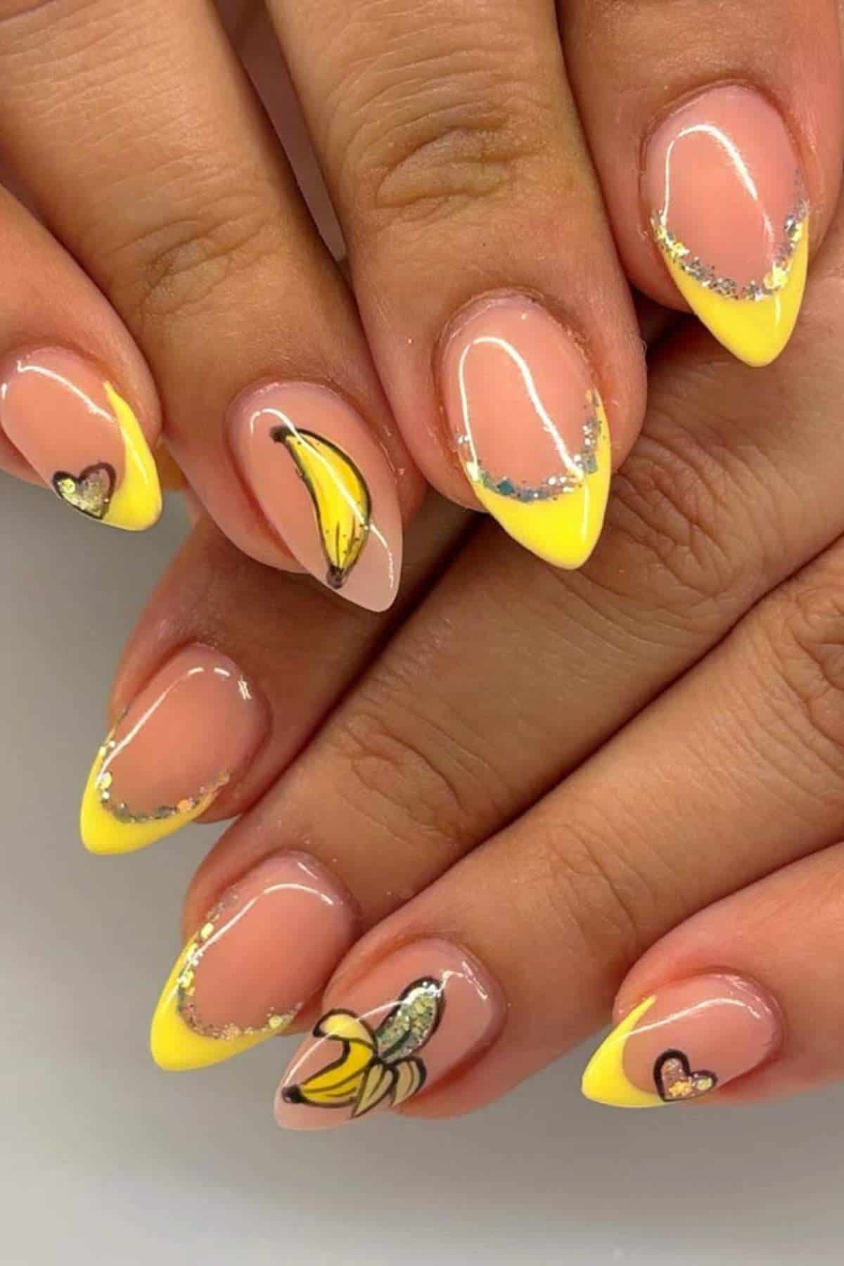 Hands with almond-shaped nails featuring yellow French tips, banana and heart designs, and silver glitter accents on a nude base.