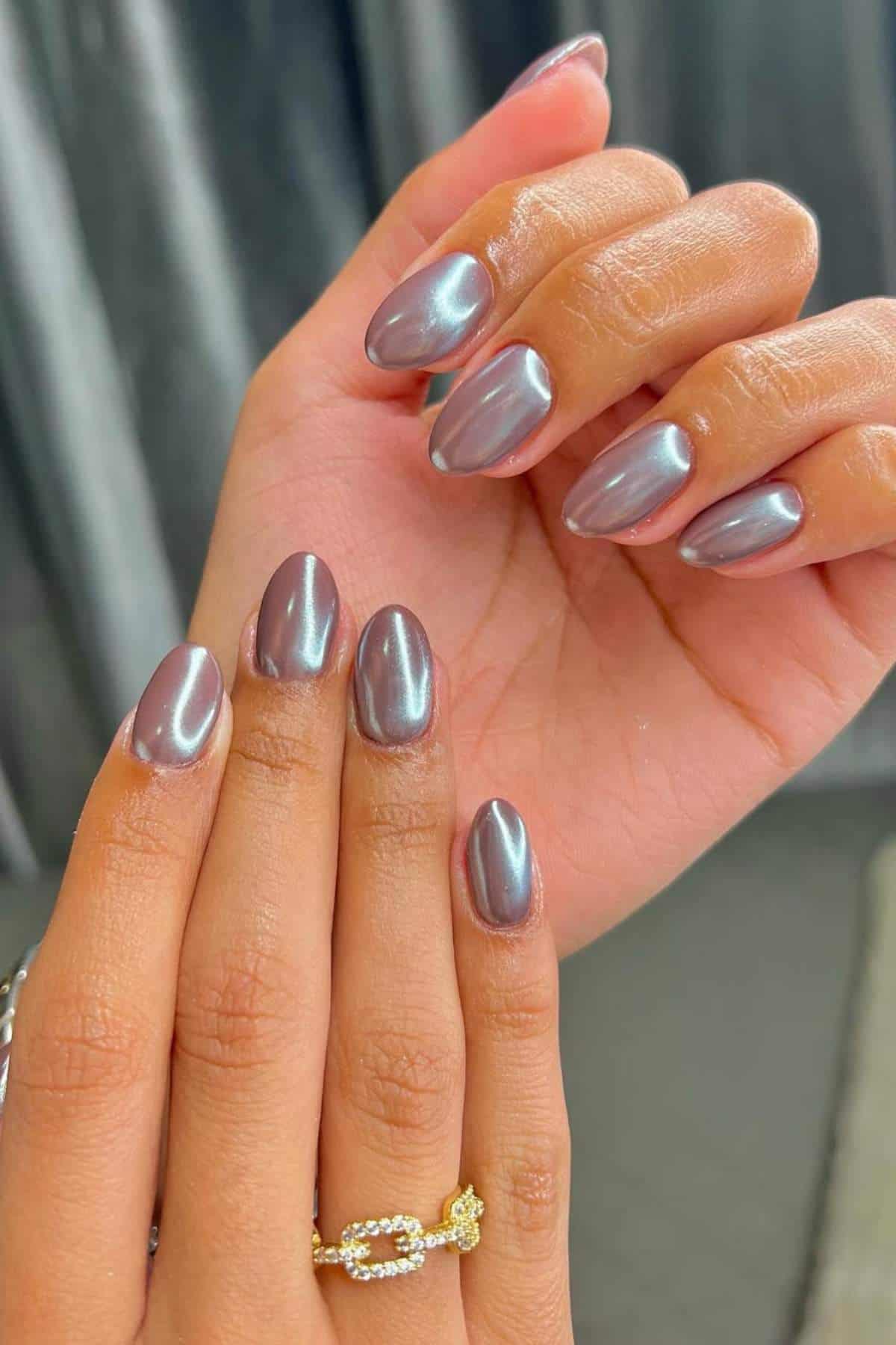 Close-up of hands with short, oval-shaped nails painted in a shiny, chrome metallic finish. One hand wears a gold ring with a chain design.