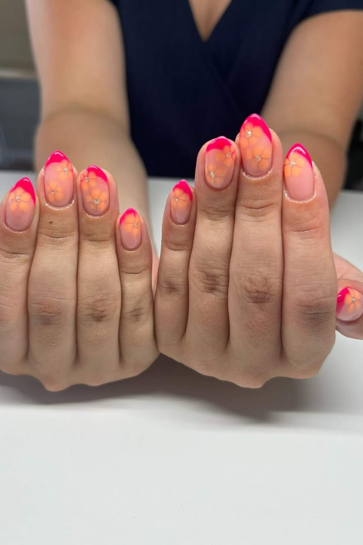 Close-up of hands showing nails with a pink and orange gradient French tip and small floral designs on each nail.
