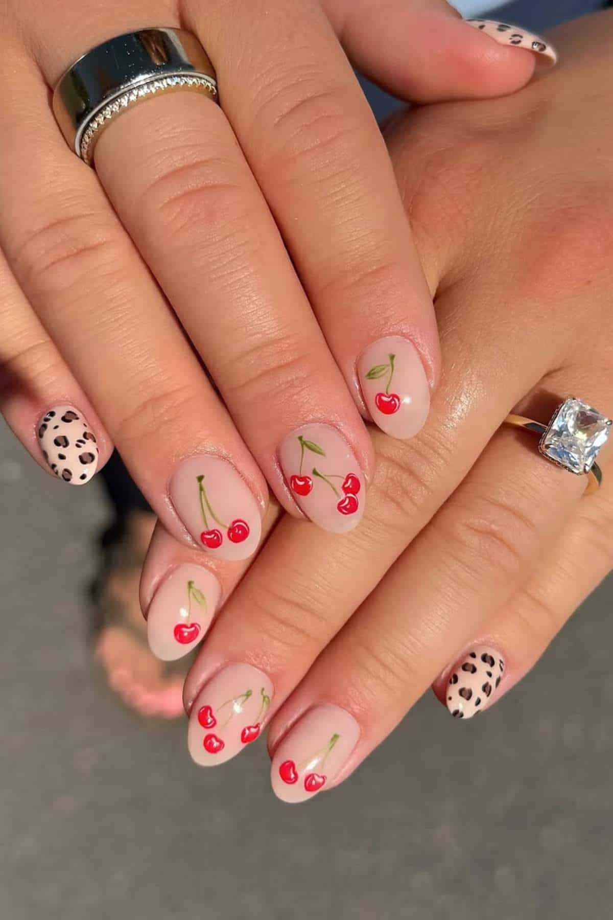 A hand with manicured nails featuring red cherry designs on most nails and a black and beige animal print pattern on the pinky fingers.
