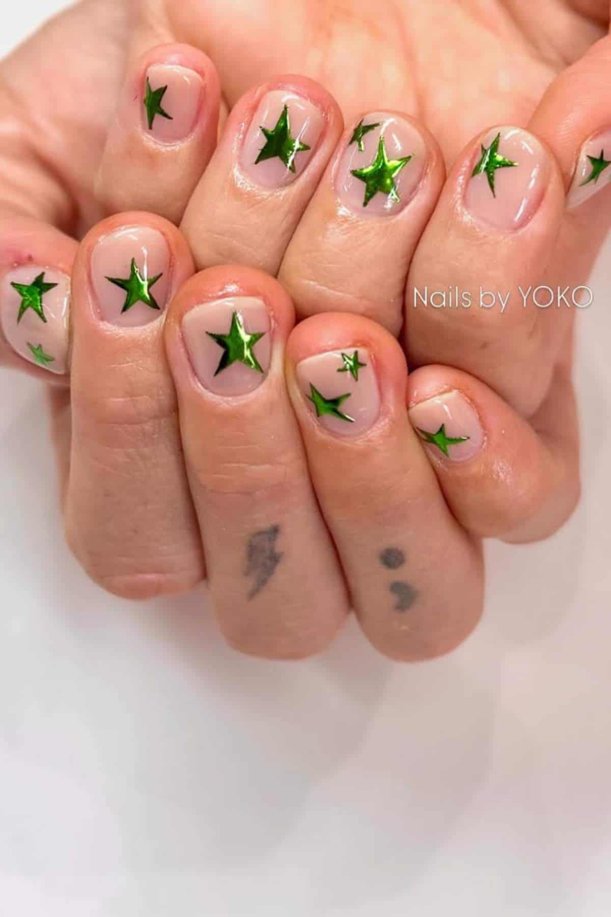 Close-up of hands with clear nail polish and green star nail art designs; tattoos of a semicolon and lightning bolt visible on fingers.