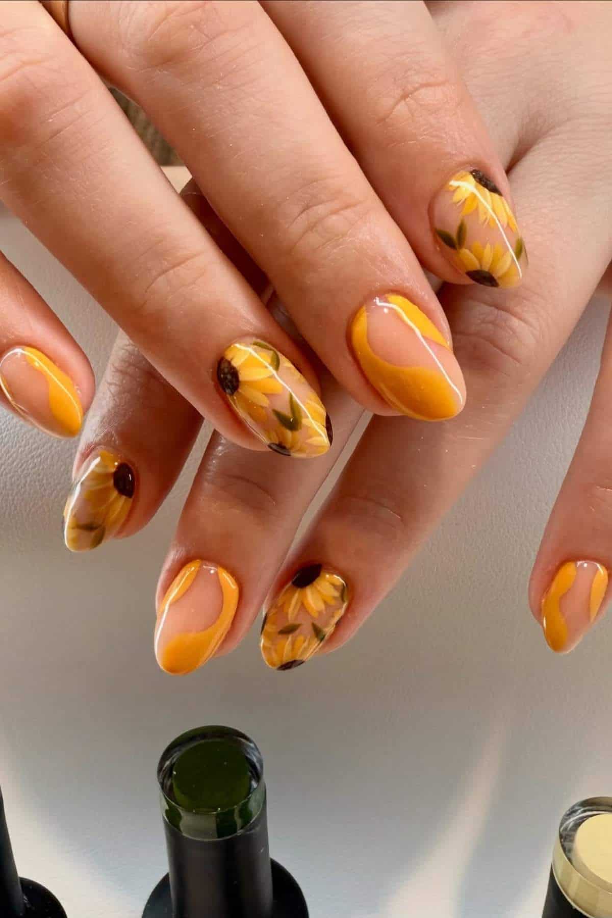Hands with almond-shaped nails painted yellow and decorated with sunflower designs, shown against a white background with nail polish bottles nearby.