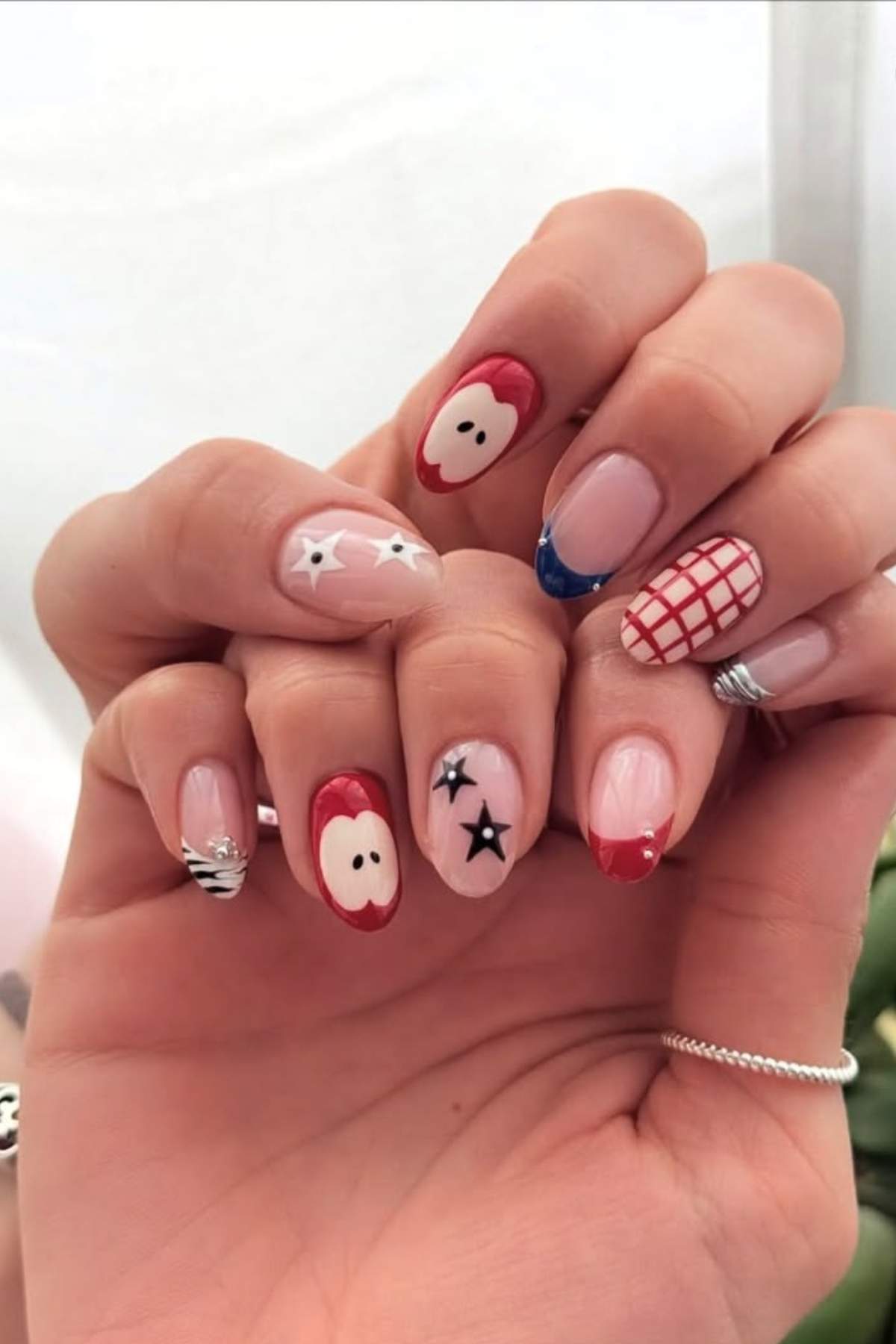 A hand with manicured nails featuring various designs, including red apples, black stars, blue tips, a red grid pattern, and silver wavy lines.