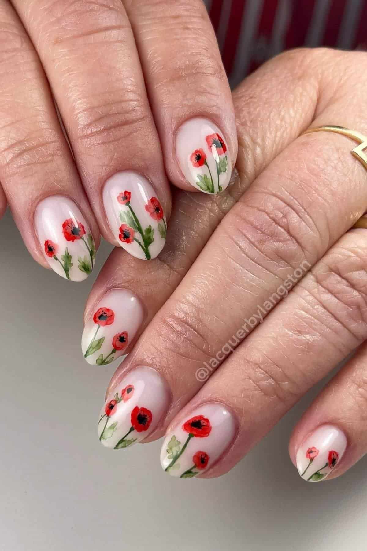 Close-up of hands with almond-shaped nails featuring a nude base and red poppy flower nail art, with green stems and leaves. The person is wearing gold rings.