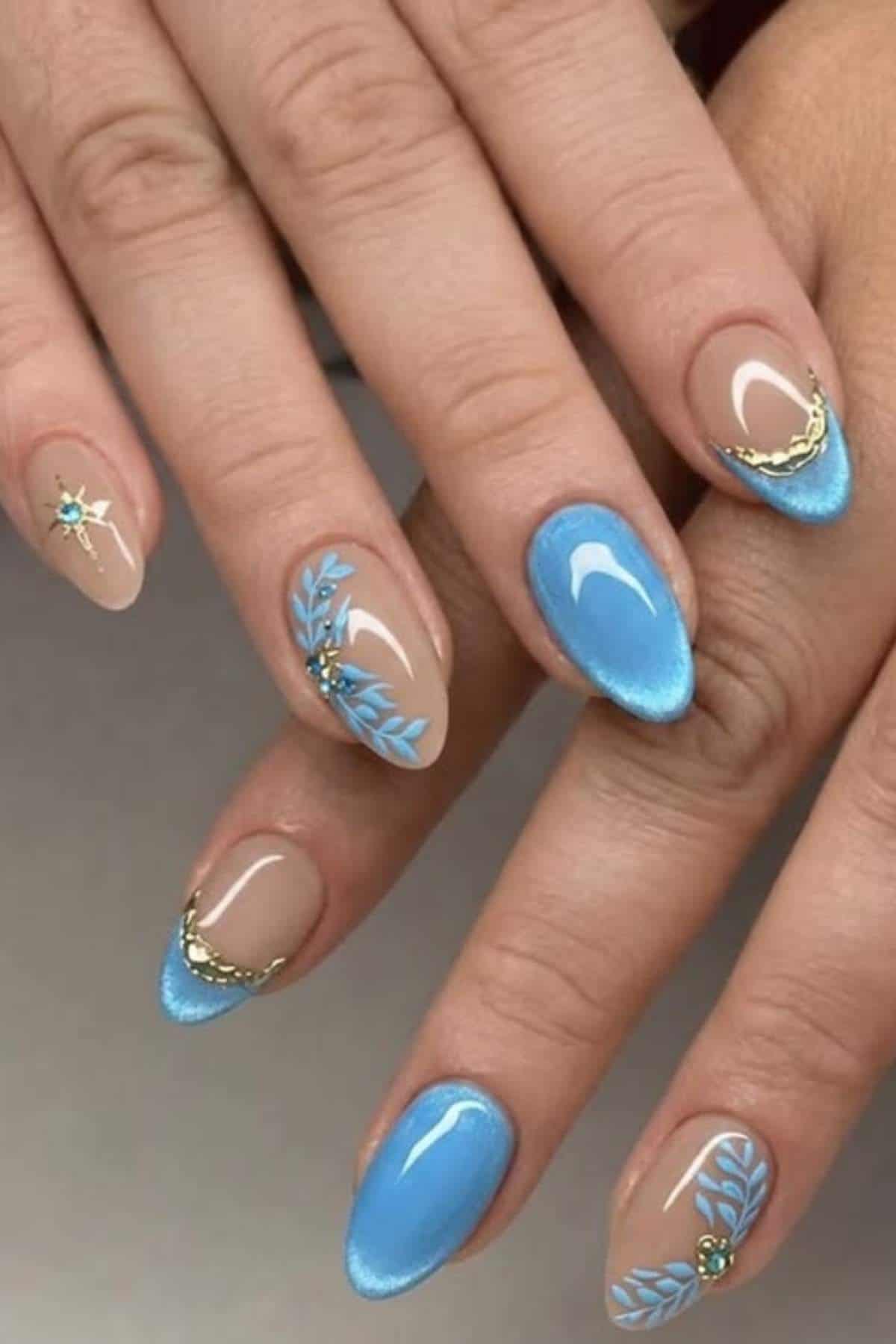 Hands with almond-shaped nails featuring a blue and nude color scheme, gold chain accents, and leaf and star designs on several nails.
