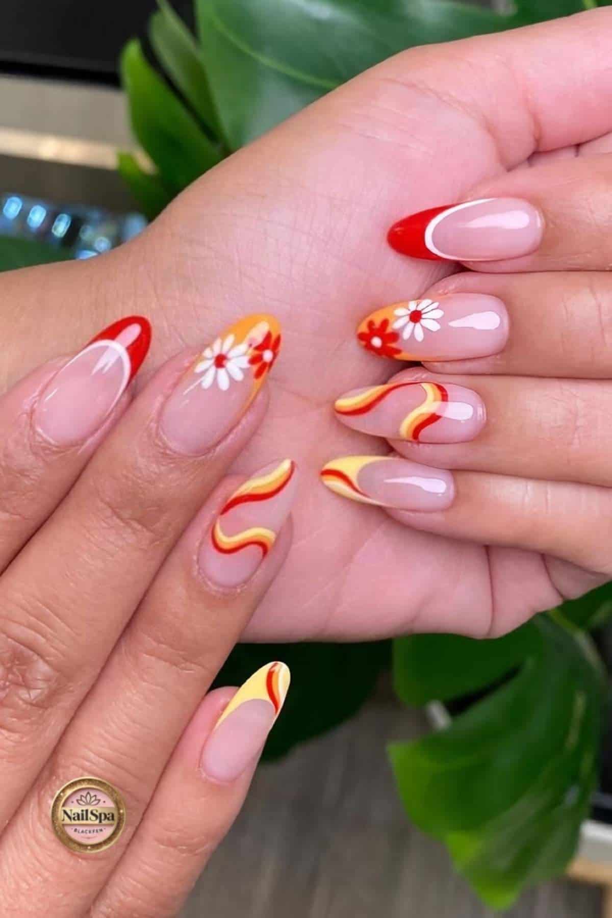 Hands with almond-shaped nails featuring red, yellow, and orange wavy designs and white daisy flowers on a nude base, with green leaves in the background.