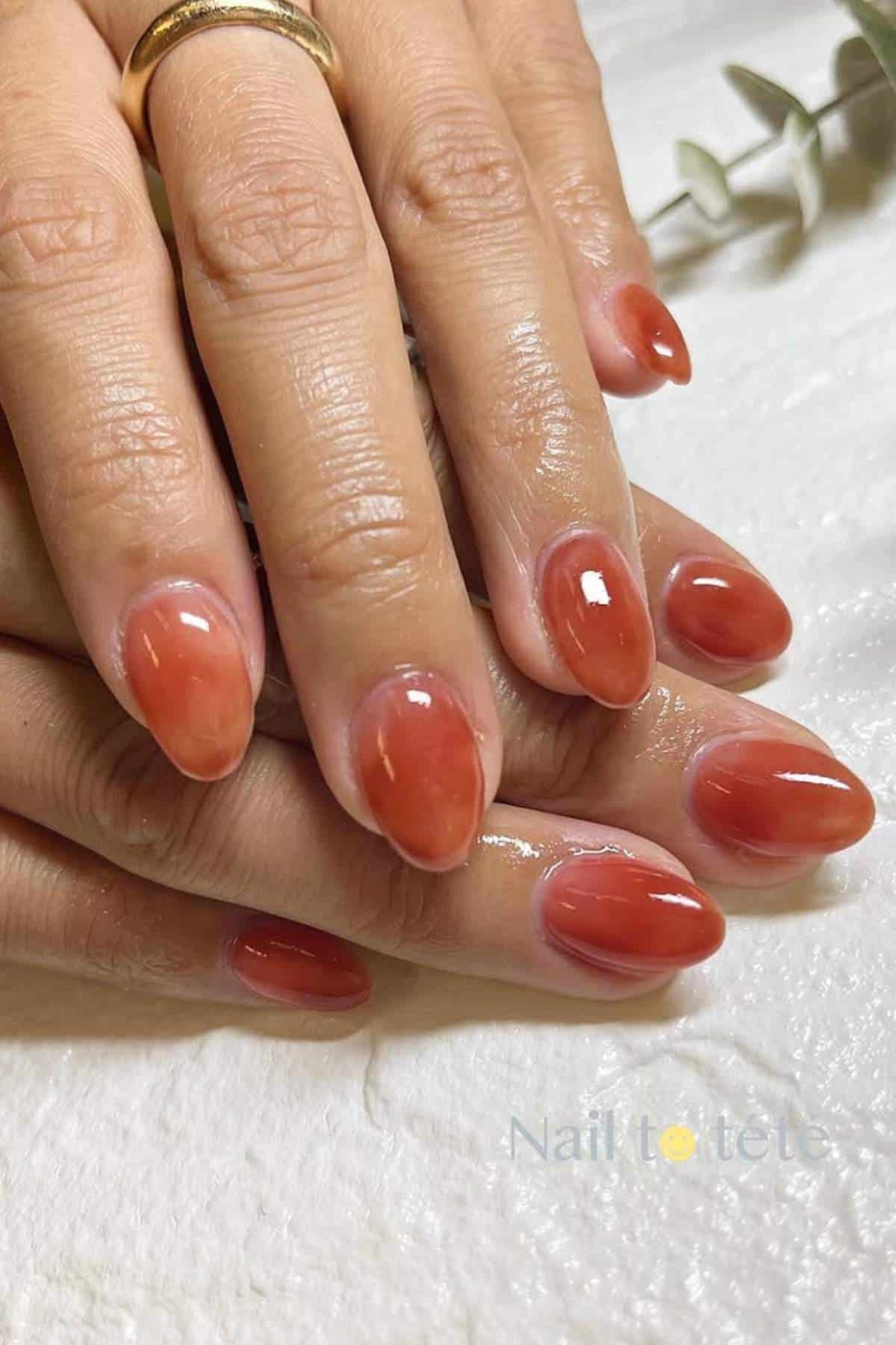Close-up of hands with almond-shaped nails painted in a glossy, reddish-brown color, resting on a white surface.