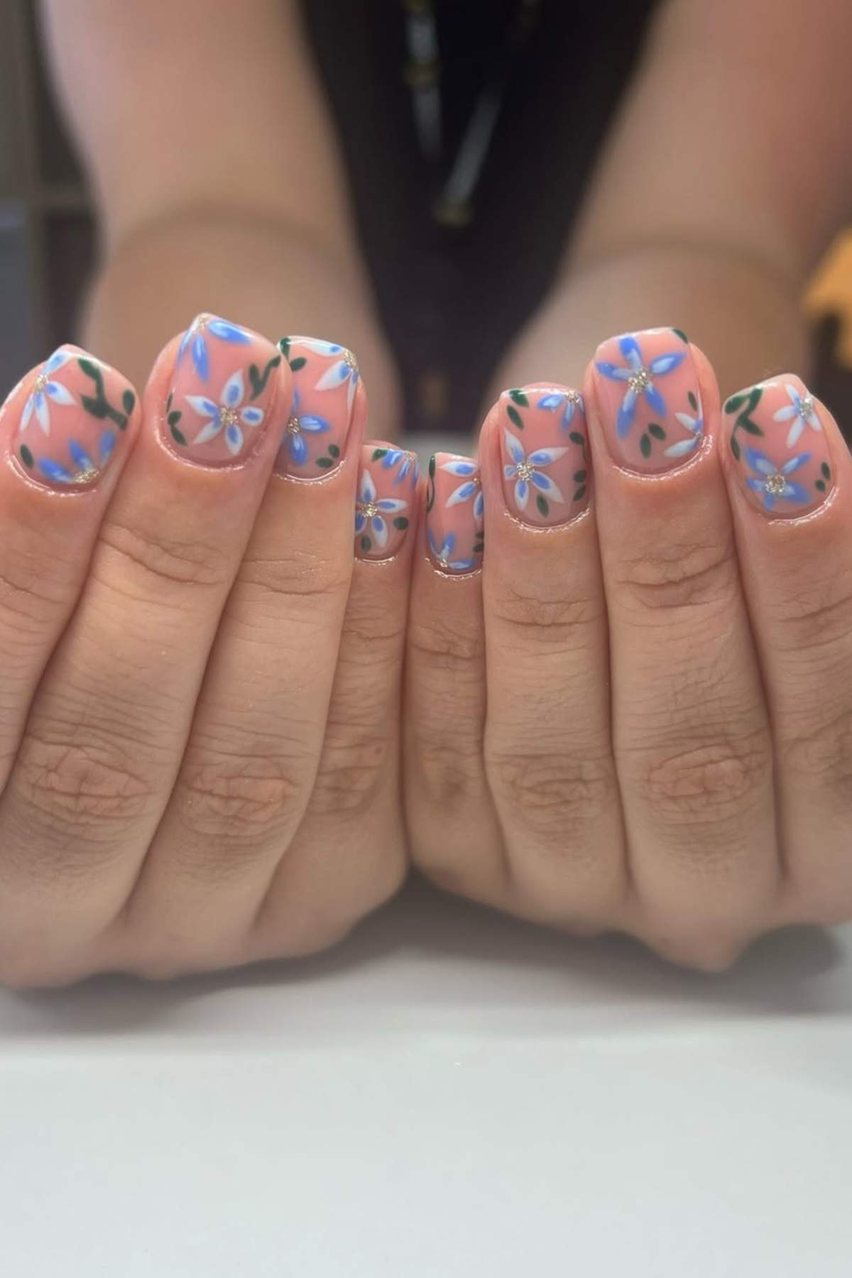 Two hands with short, rounded nails featuring a nude base color and blue and purple floral designs with green leaves on each nail.