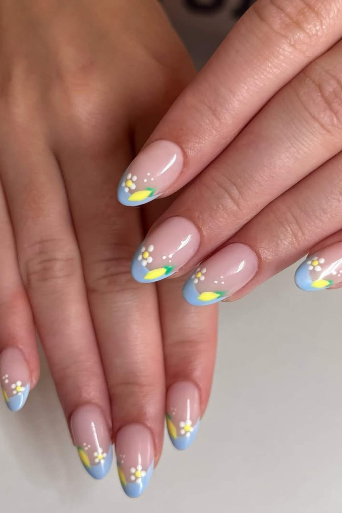 A hand with almond-shaped nails featuring light blue tips and small white and yellow floral designs on a natural pink base.