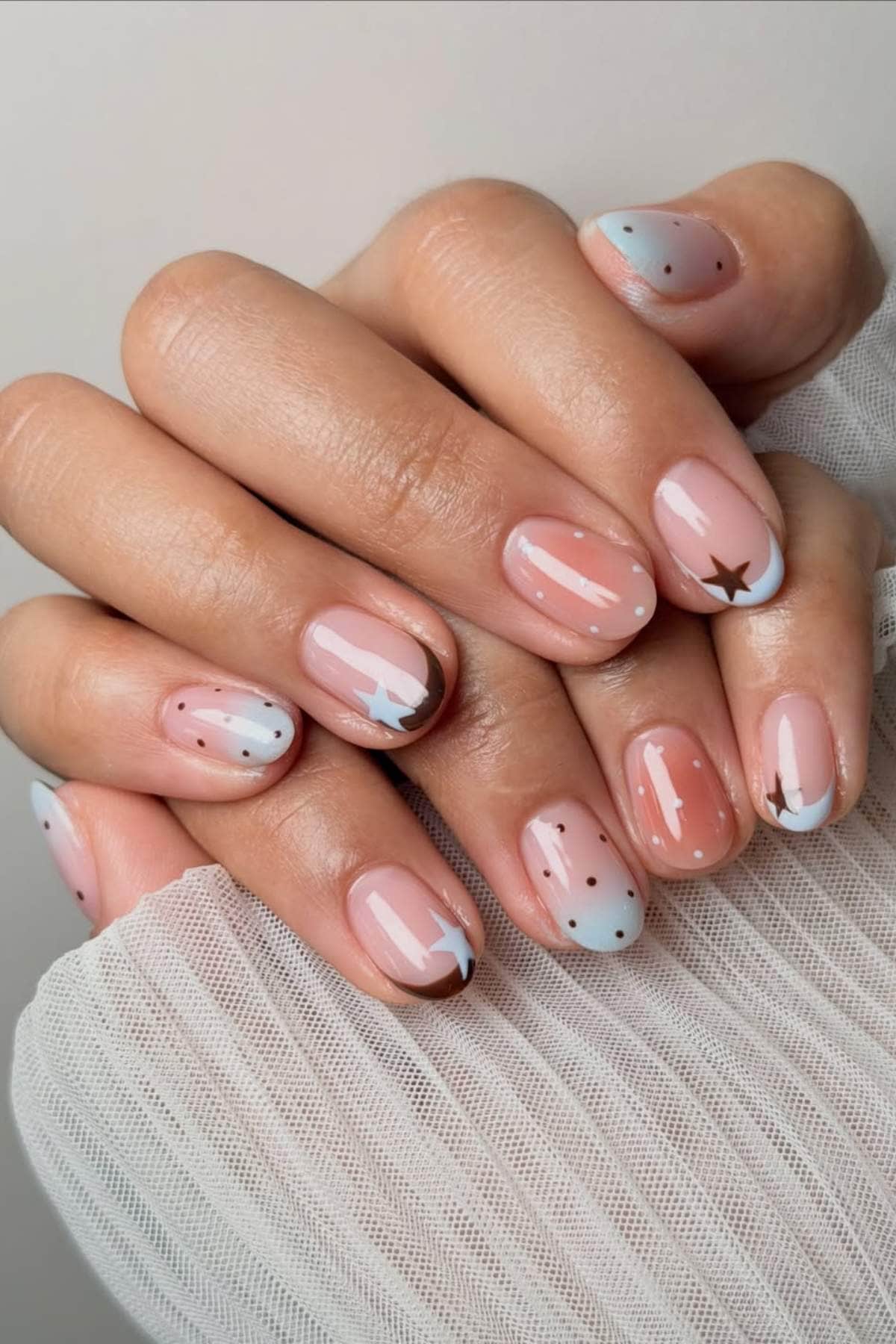 A close-up of hands with nude and blue gradient manicured nails, decorated with small white dots, stars, and brown star accents.