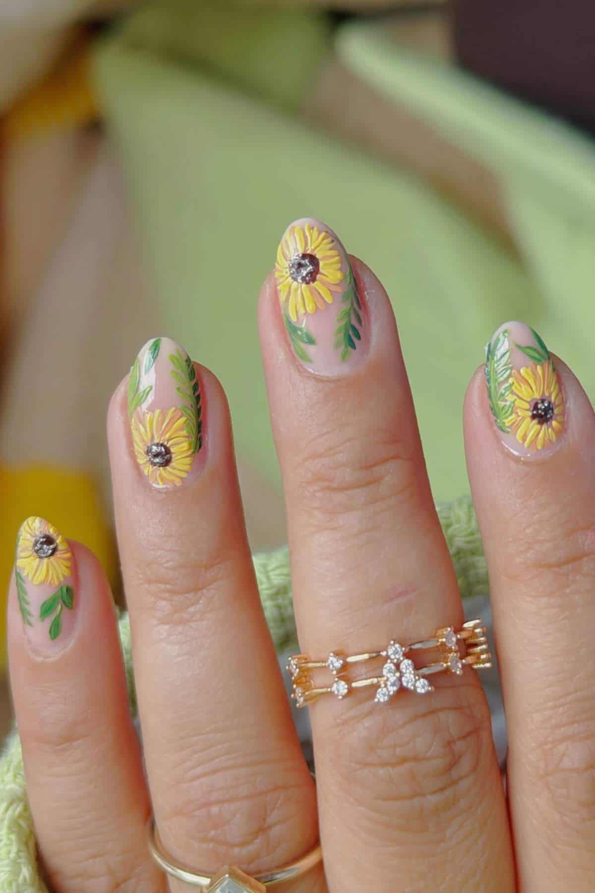 A hand with manicured nails featuring yellow sunflower designs and green leaves, wearing gold and diamond rings.