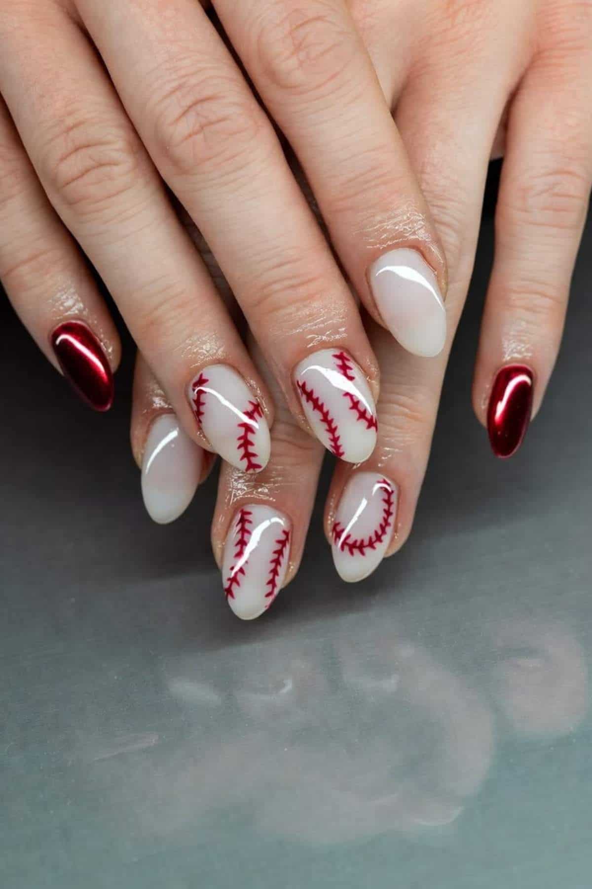 Close-up of hands with manicure featuring baseball stitching designs on two nails, plain white polish on one, and metallic red polish on the others.