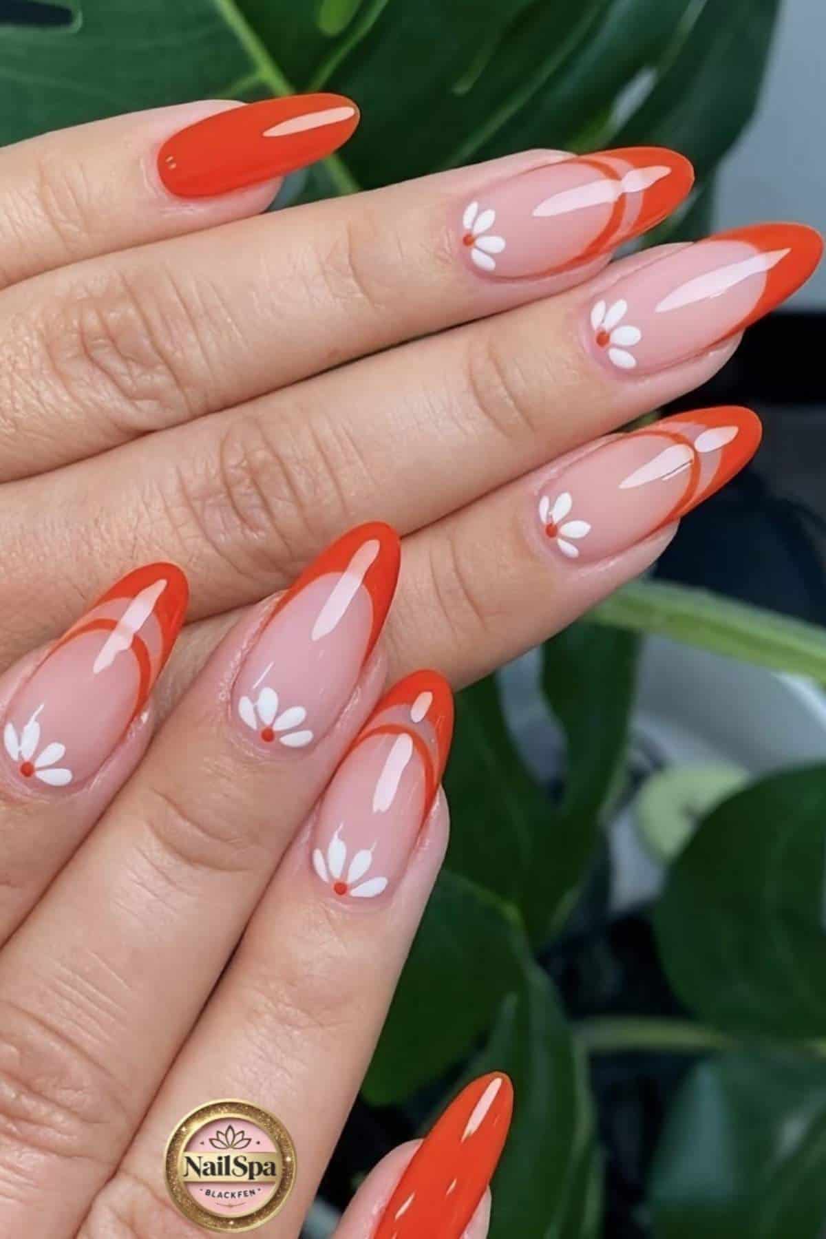 A hand with almond-shaped nails featuring red French tips and small white daisy designs on a nude base.