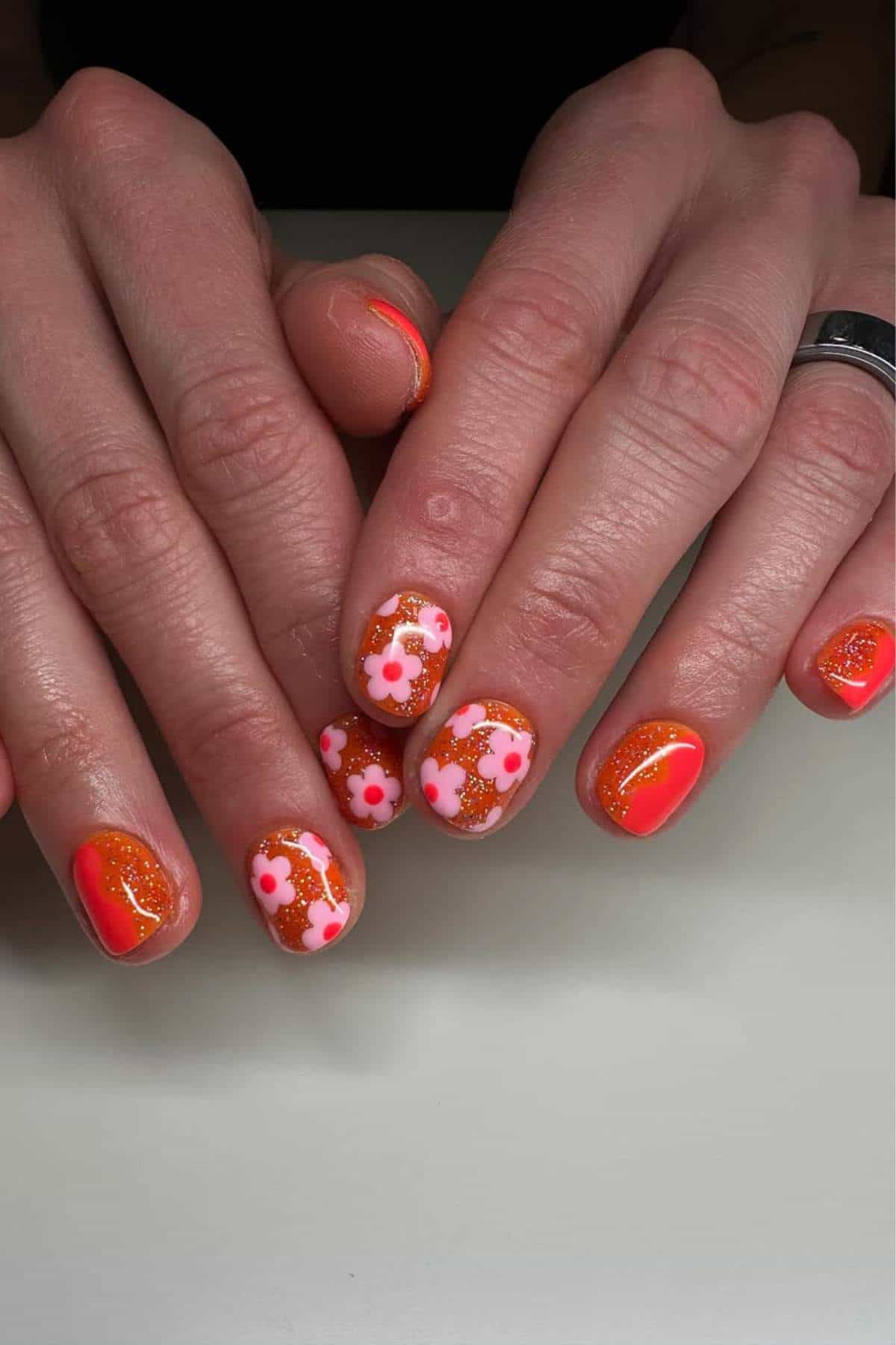 Hands with short nails featuring orange and pink nail polish; some nails have floral designs and glitter accents.