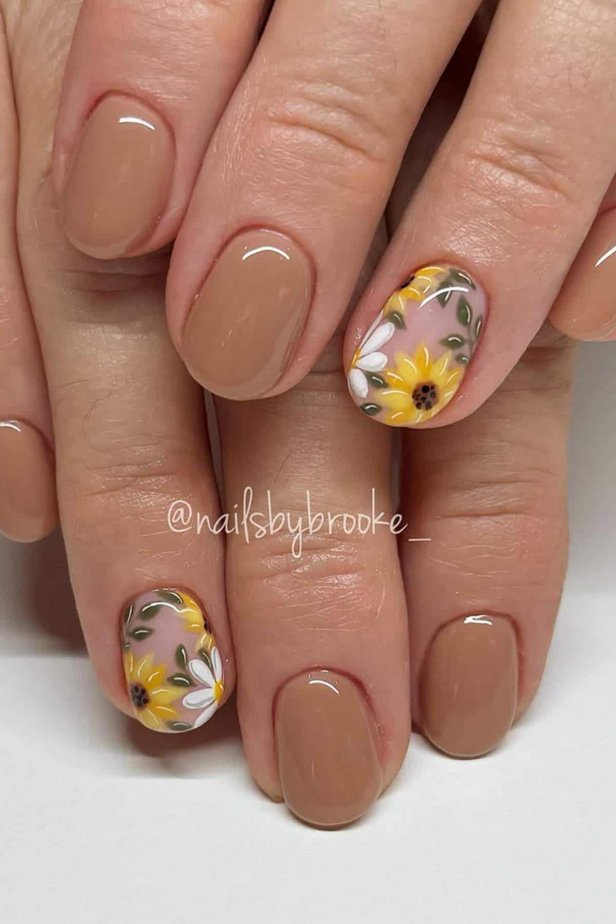 A hand with short, oval nails painted nude; the ring and thumb nails feature yellow sunflower and white daisy floral nail art.