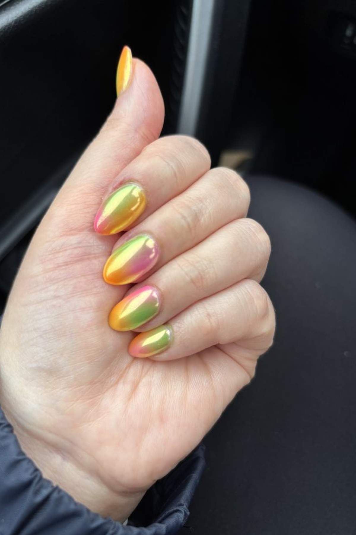 A hand with almond-shaped nails featuring a shiny, metallic gradient in yellow, green, and pink hues.