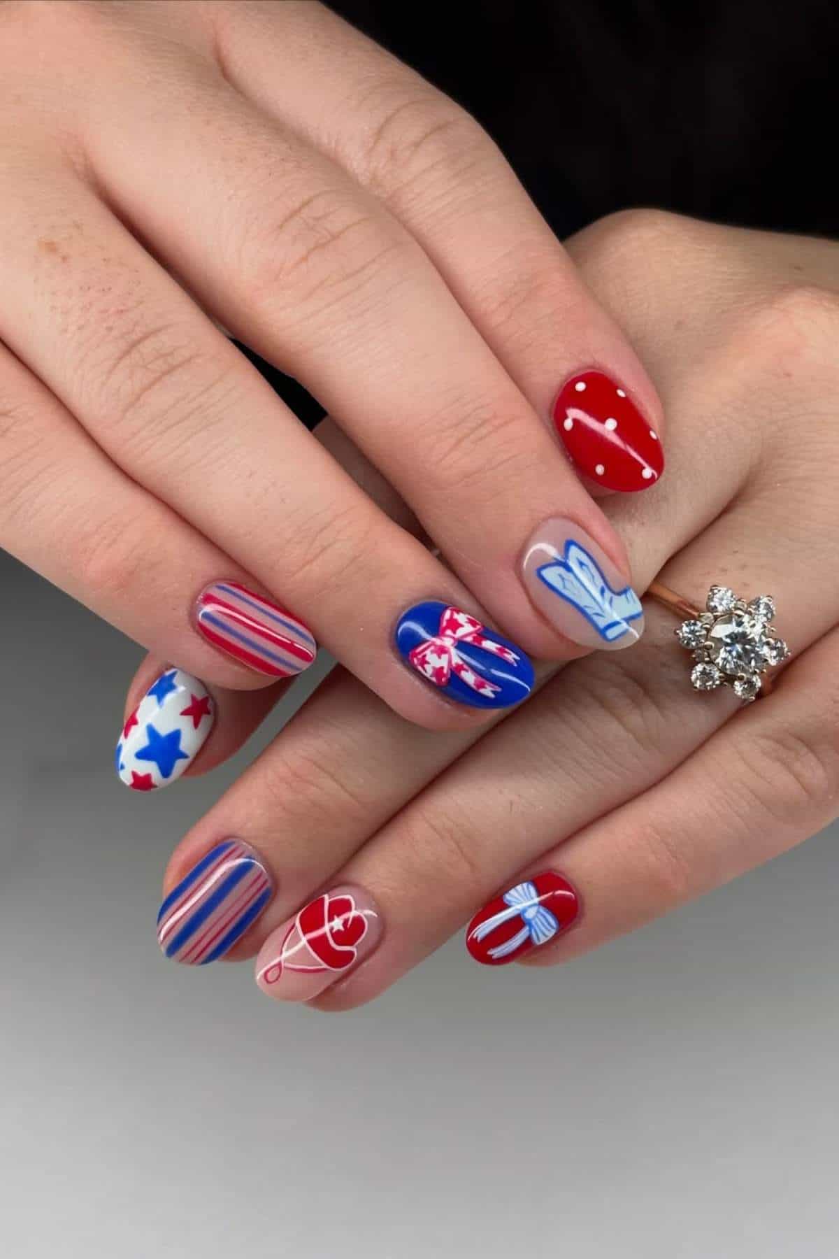 Hands with manicured nails featuring red, white, and blue designs, including stars, stripes, hearts, and bows; one hand wears a floral-shaped ring.