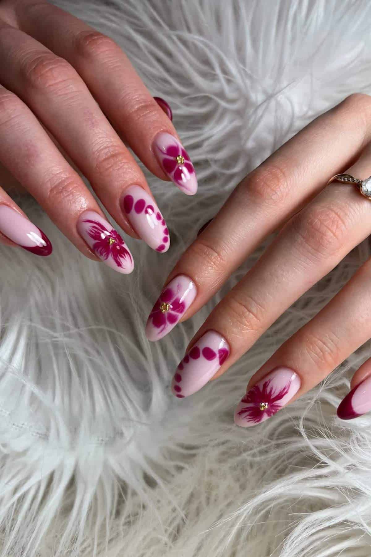 Hands with long, almond-shaped nails featuring pink and burgundy floral nail art, dots, and French tips, displayed on a white, fluffy surface.