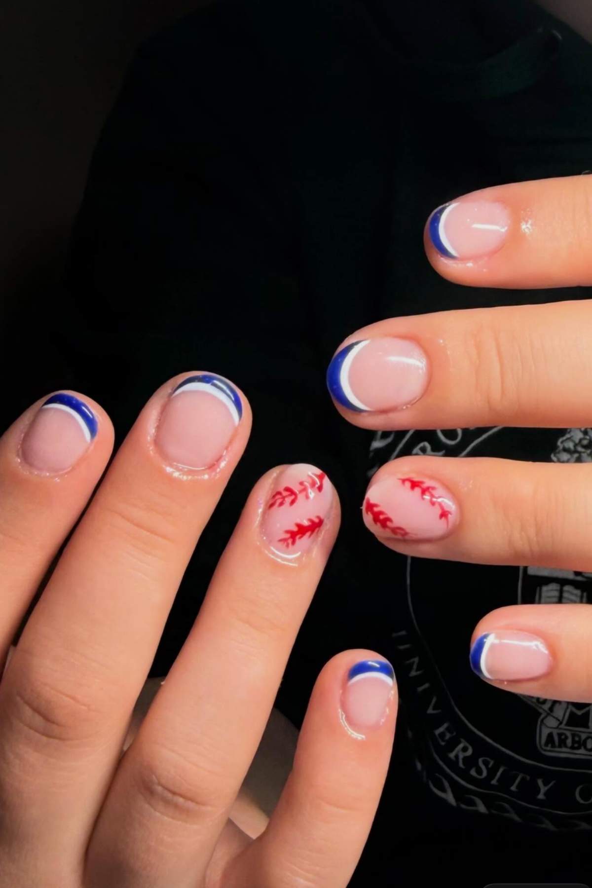 Hands with short nails; most nails have blue and white French tips, while two nails feature red baseball stitching designs on a nude base.