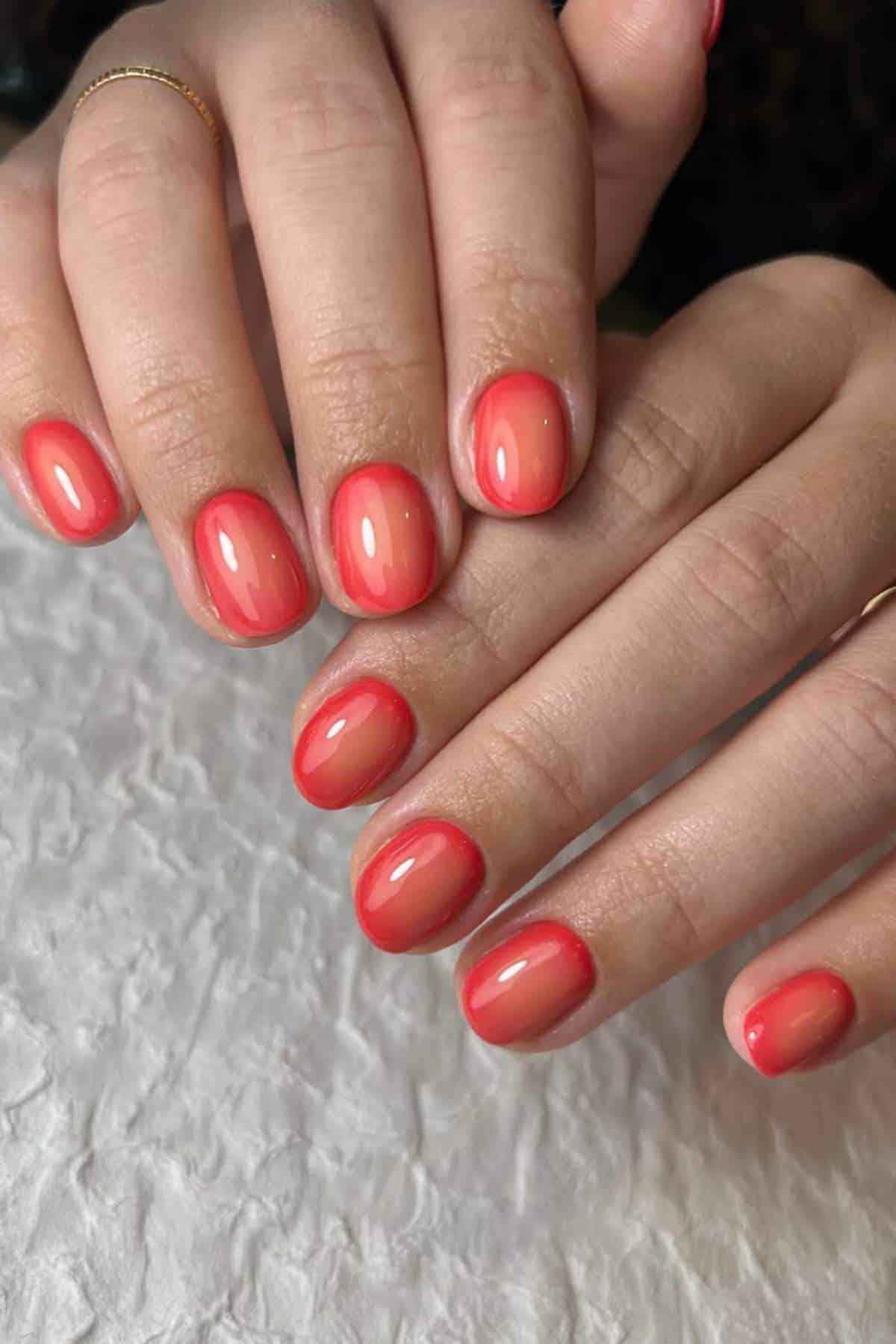 Close-up of hands with short, rounded nails painted in glossy coral-red nail polish, resting on a textured white surface.