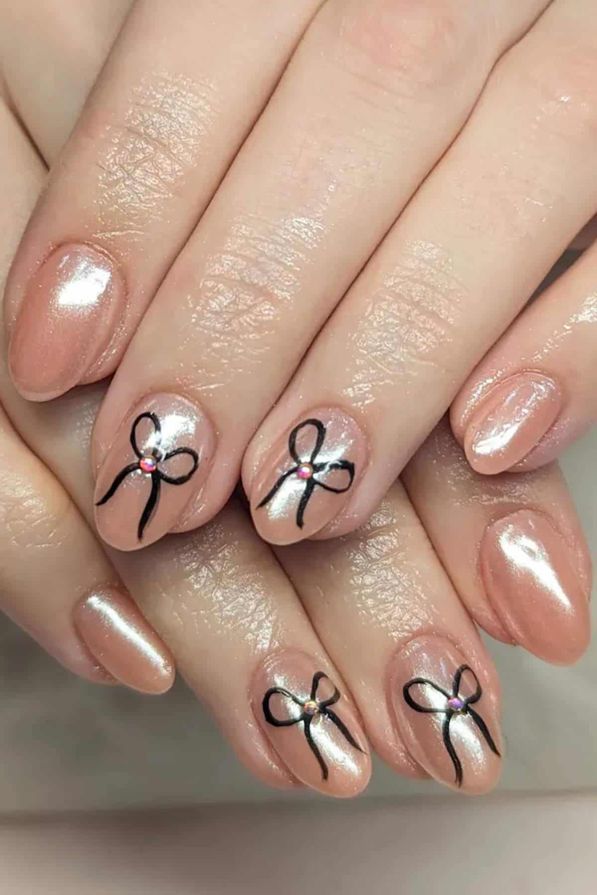 Close-up of hands with pearly nude nail polish; some nails feature black bow designs with a small rhinestone at the center.