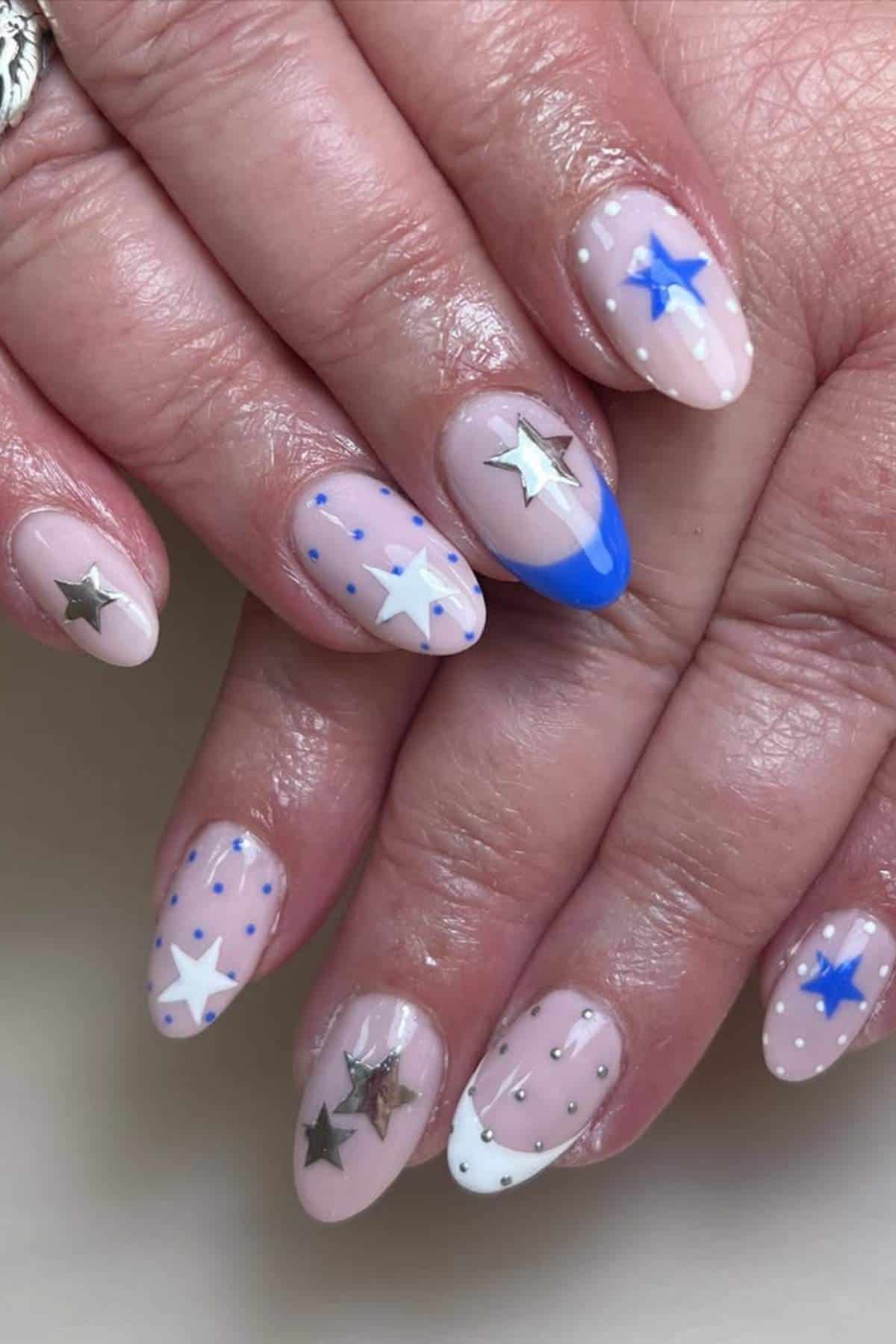 Hands with almond-shaped nails featuring nude polish, blue and white stars, silver star decals, and small blue dot accents.