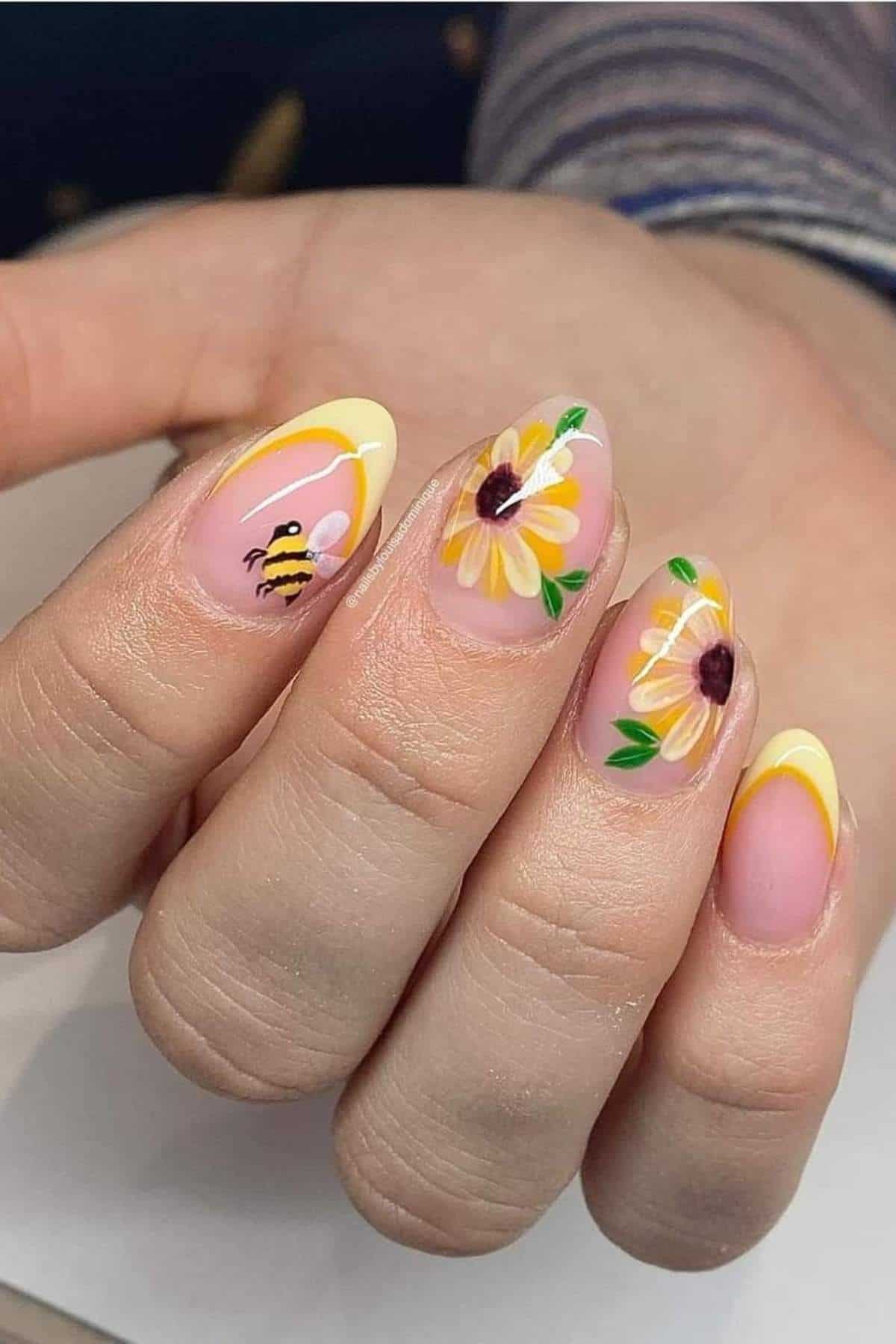 A hand with almond-shaped nails painted with yellow tips, daisy flower designs, and a small bee illustration on a nude base.