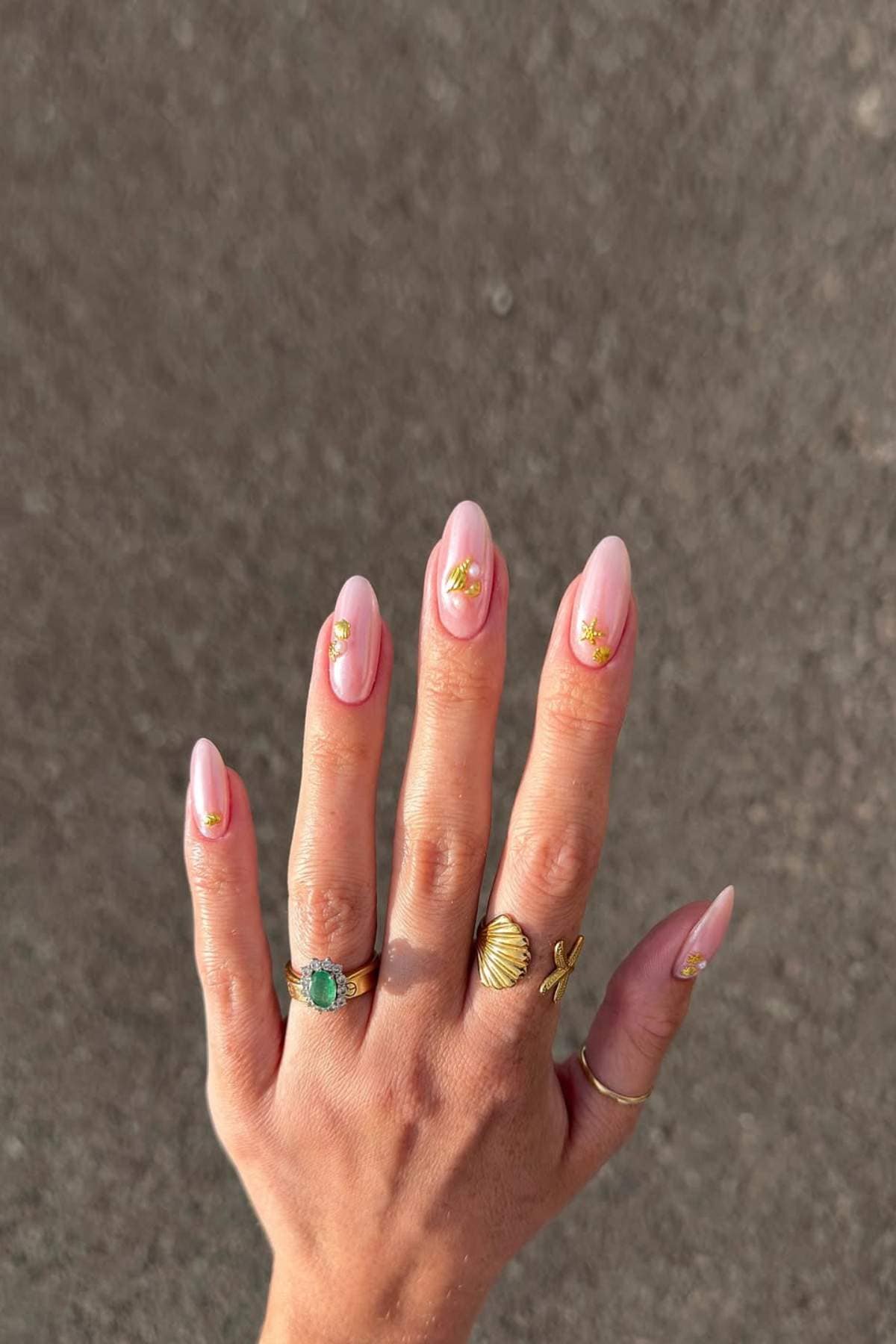 A hand with almond-shaped, light pink manicured nails featuring small gold embellishments, wearing three gold rings and one green stone ring against a neutral background.