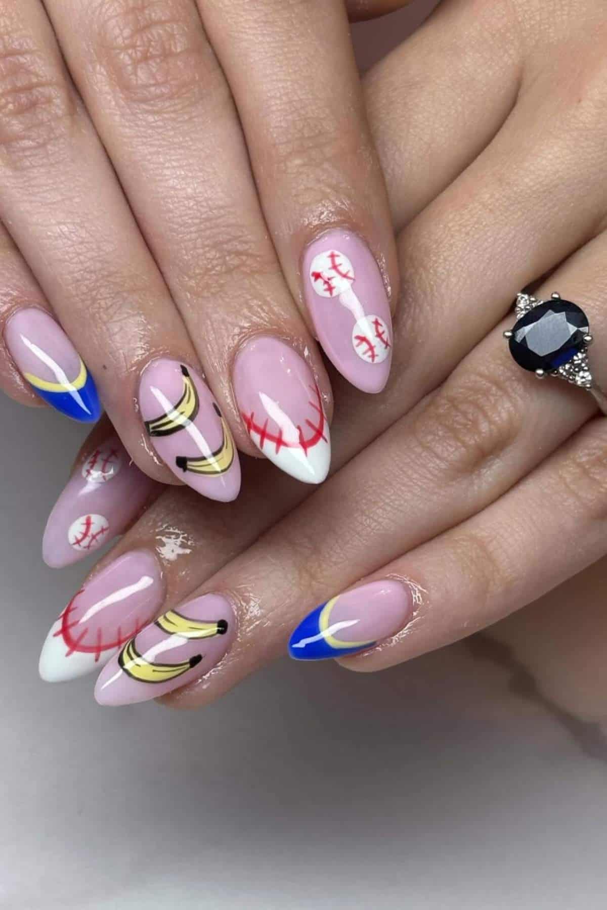 Hands with almond-shaped nails featuring pink polish, baseball stitching designs, gold and black abstract lines, and blue tips; one hand wears a dark gemstone ring.