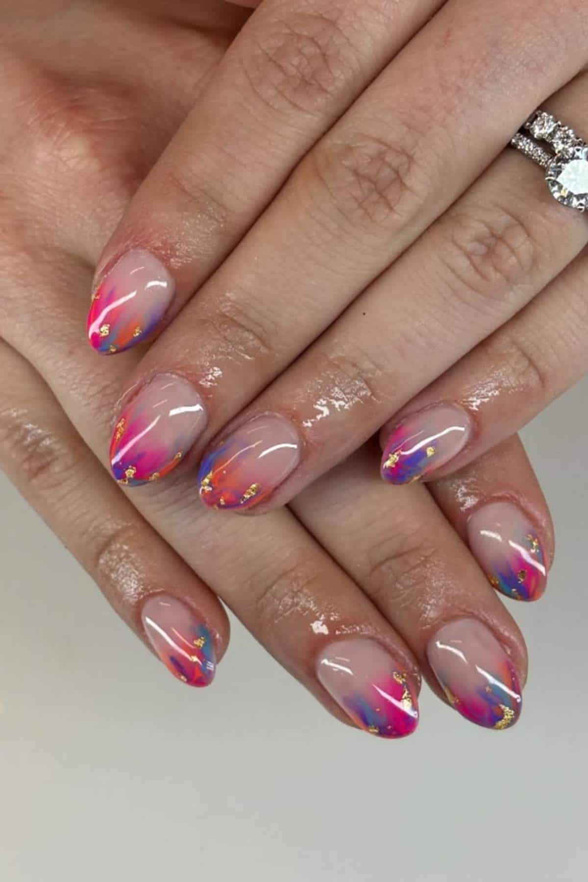 Hands with almond-shaped nails featuring a colorful abstract design with pink, blue, orange, and gold accents on a nude base. A diamond ring is worn on one finger.