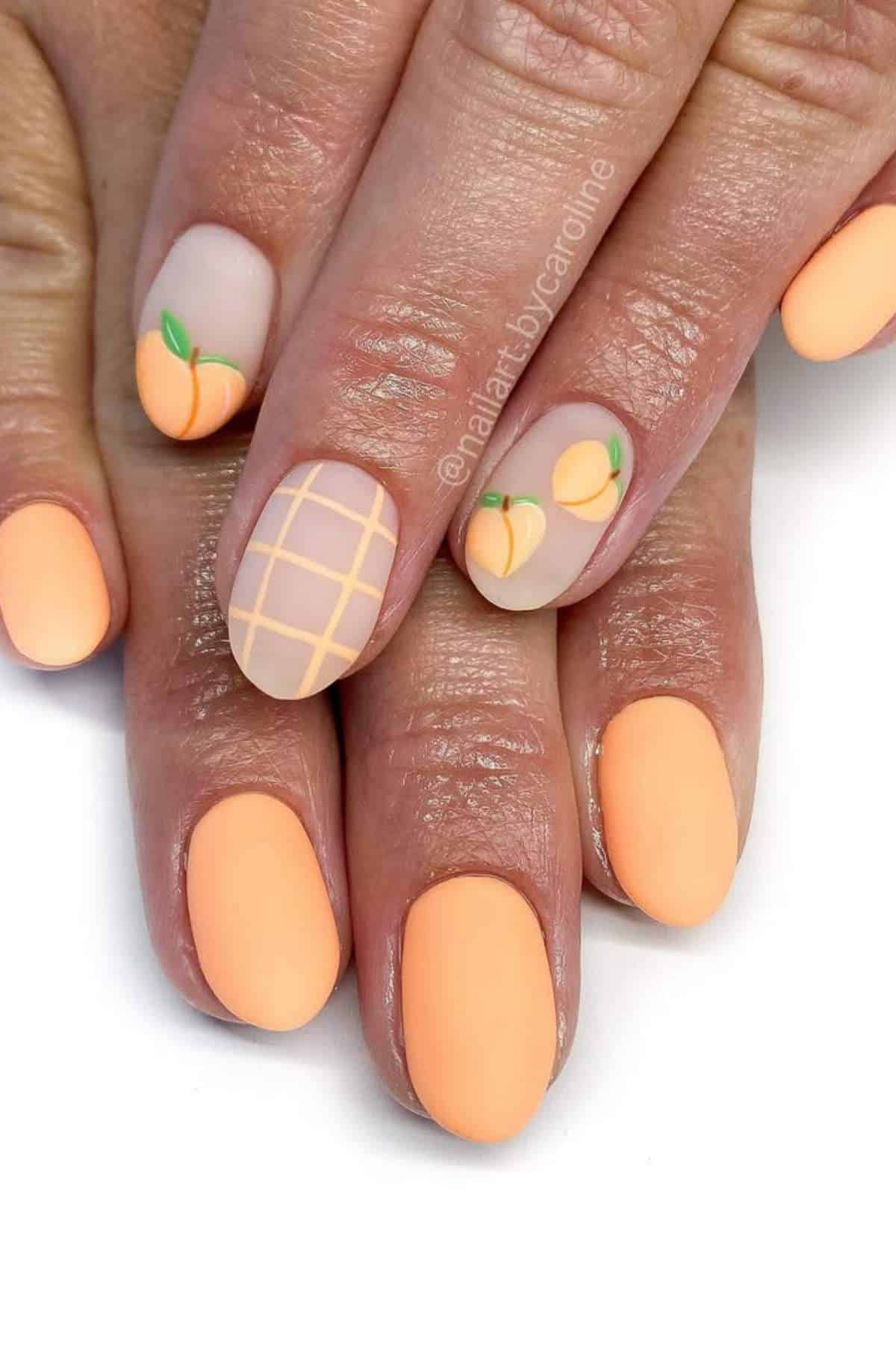 Hands with matte peach-colored manicured nails, featuring small peach fruit designs and a grid pattern on two accent nails.
