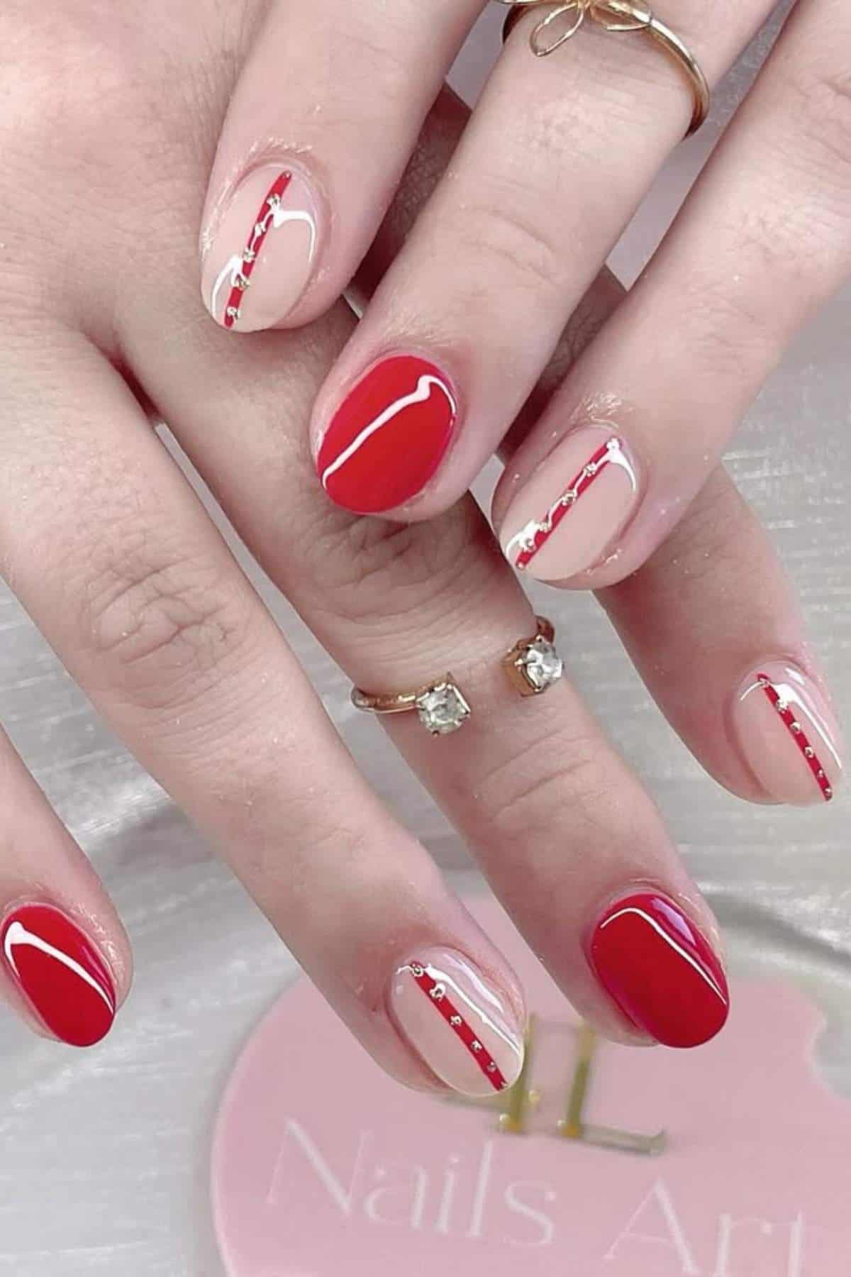Hands with manicured nails featuring a red and nude color block design, accented with thin red and silver glitter lines.