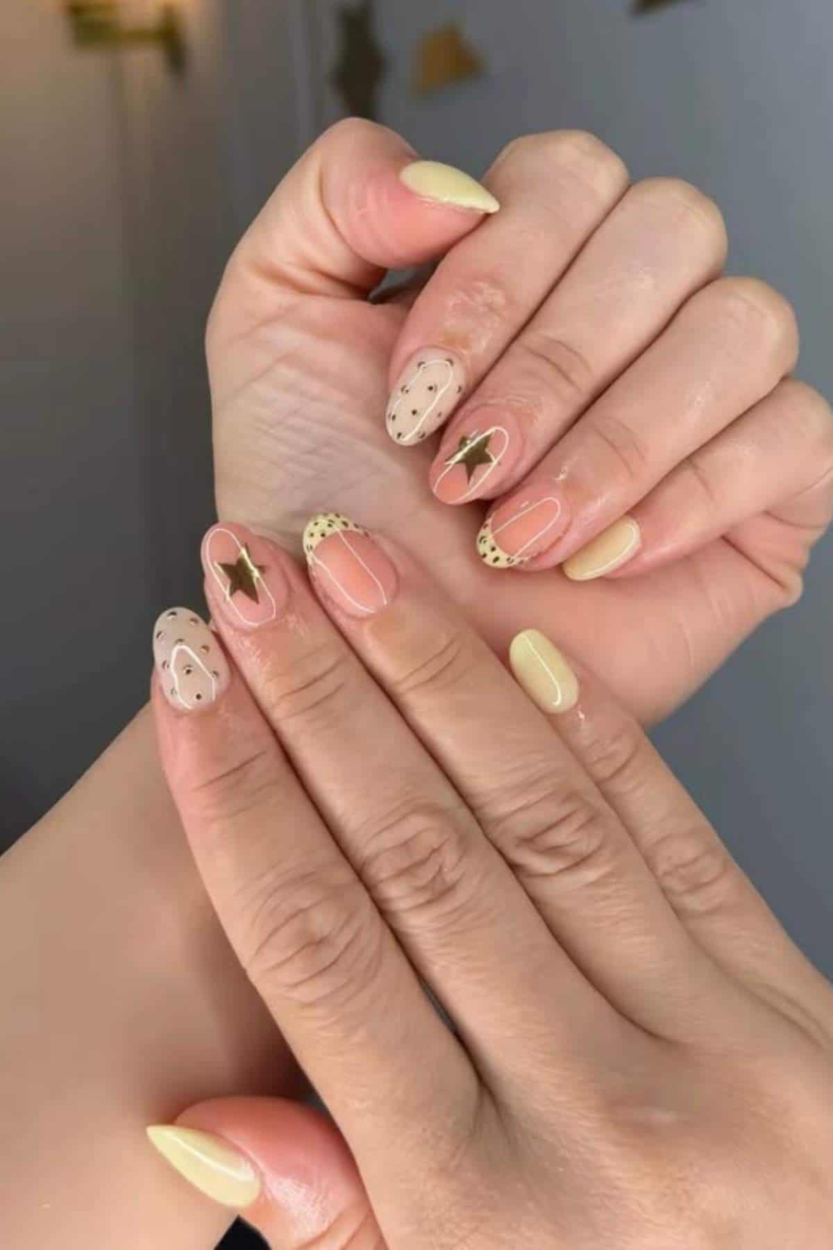 Hands with almond-shaped nails painted in pale yellow and clear polish, featuring gold star accents and black dotted designs on some nails.