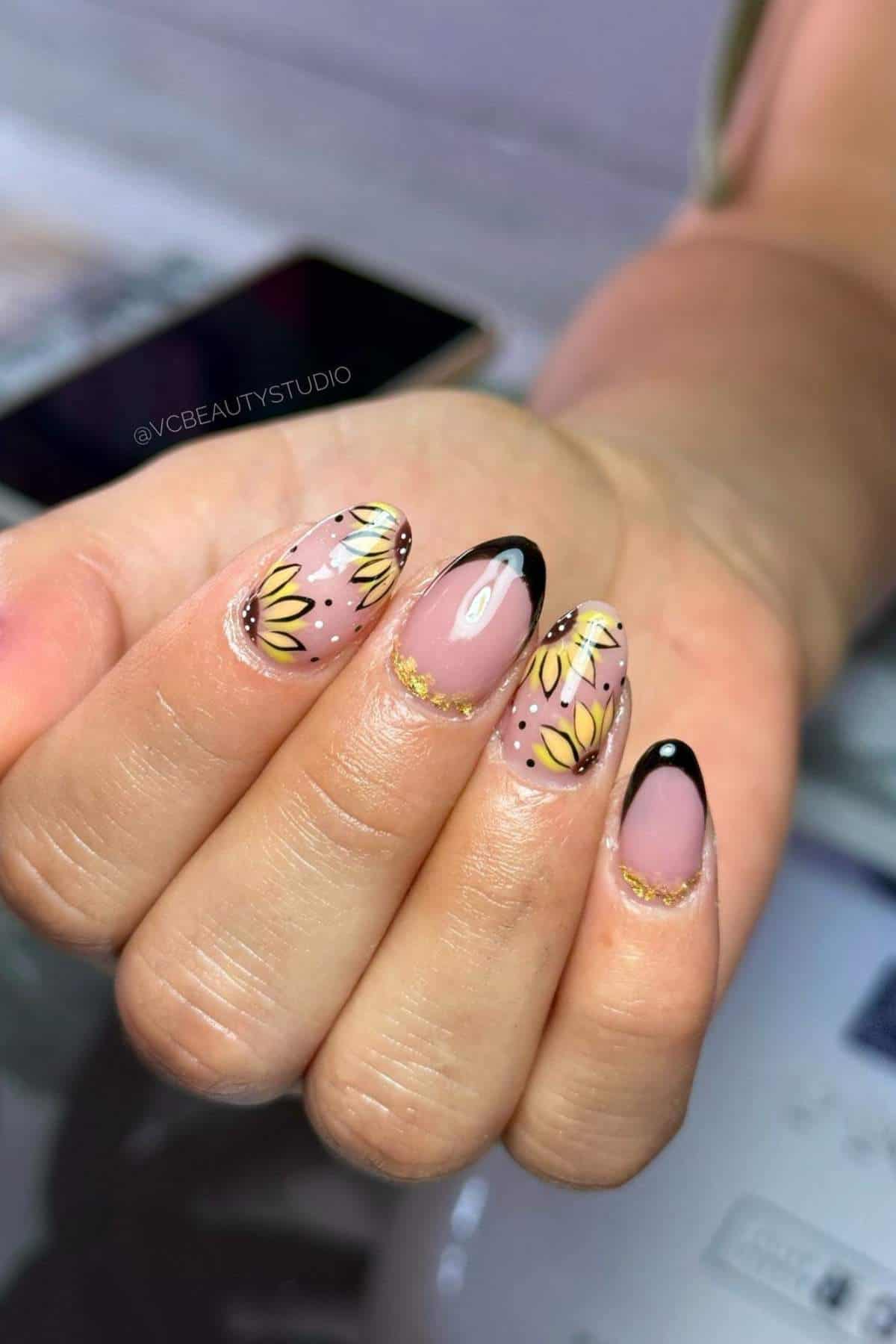 A hand with almond-shaped nails featuring sunflower designs, black French tips, and gold accents, holding a smartphone in the background.