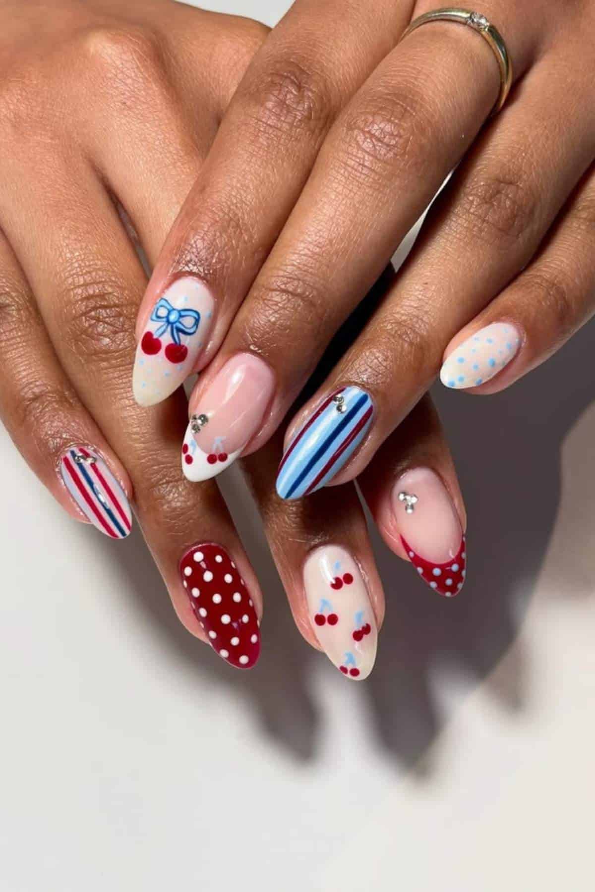 Close-up of hands with almond-shaped nails, decorated in red, blue, and white with cherries, stripes, polka dots, and a small blue bow design.