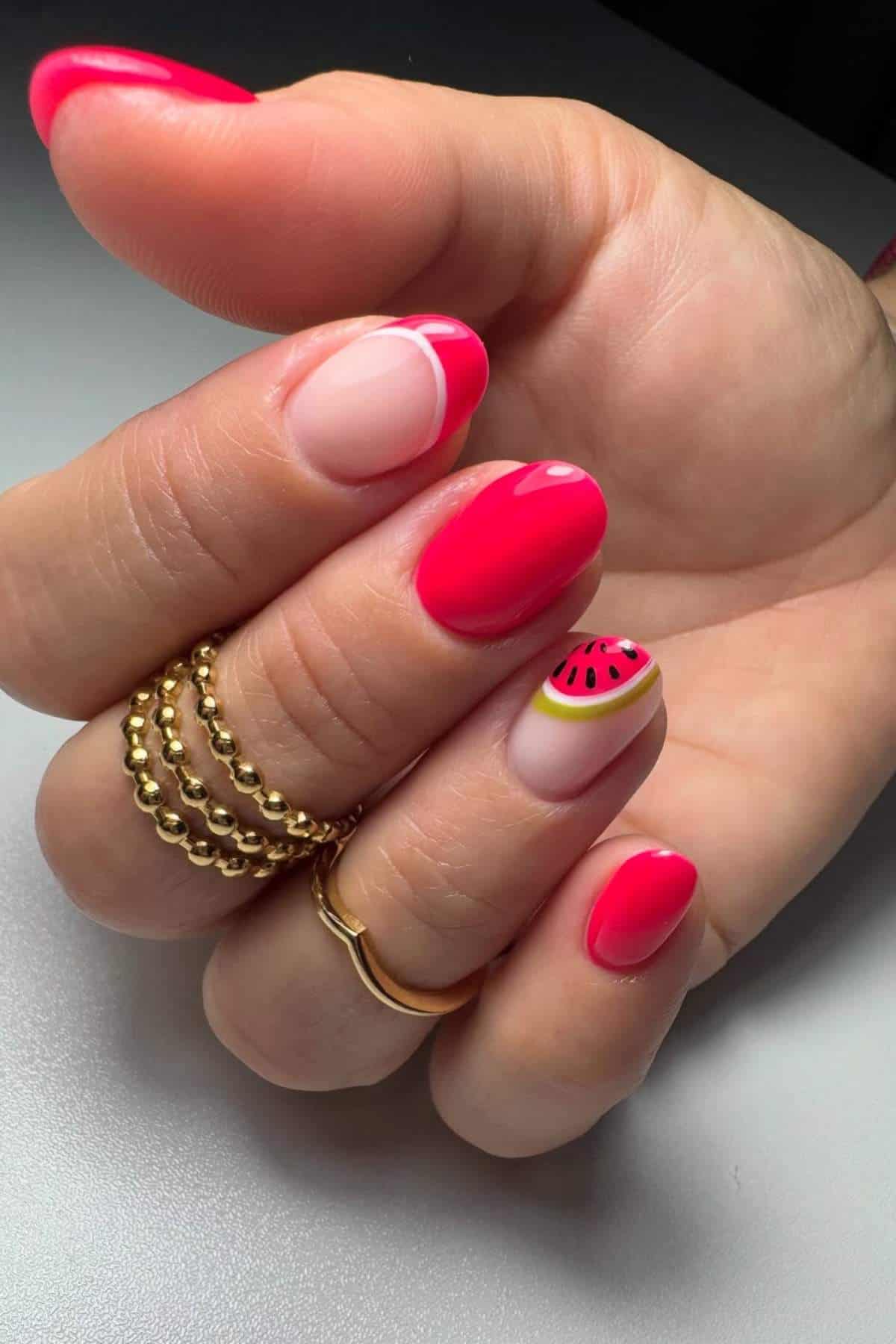 A hand with gold rings displays bright pink manicured nails, one featuring a watermelon design and another with a minimalist nude and pink curve accent.