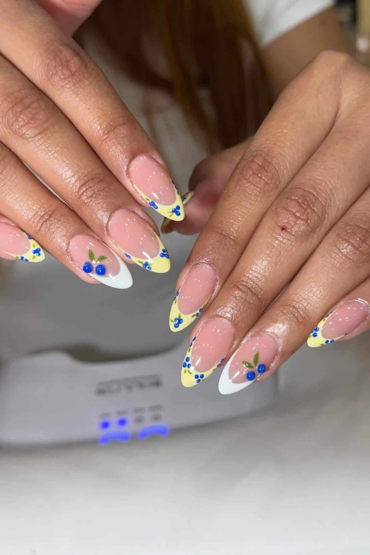 Hands with almond-shaped nails featuring a yellow French tip, small blue and yellow flower designs, and blue rhinestone accents. A white nail lamp is in the background.