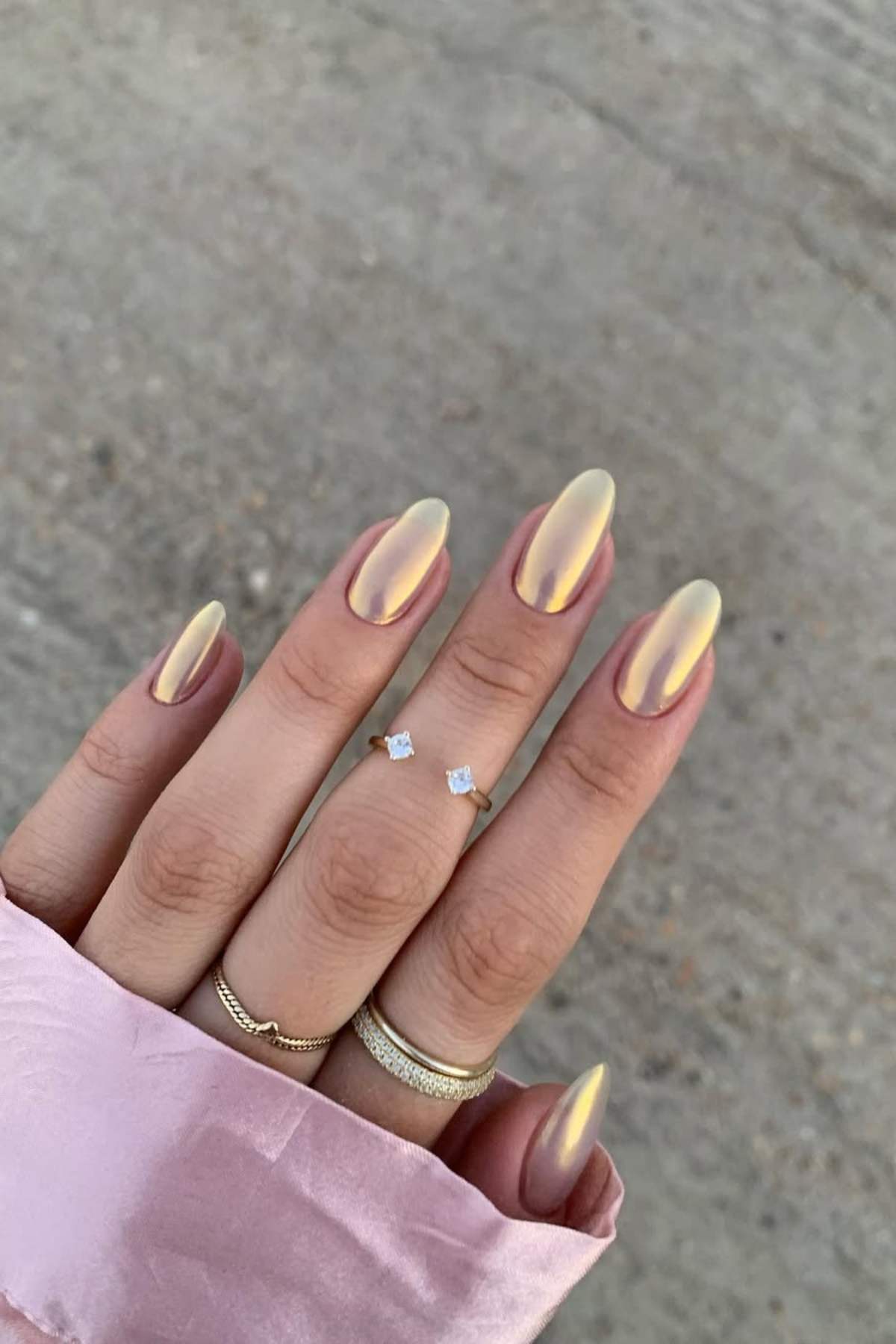 A hand with long, almond-shaped nails painted in a shimmery gold polish, wearing three gold rings and a pink satin sleeve visible.
