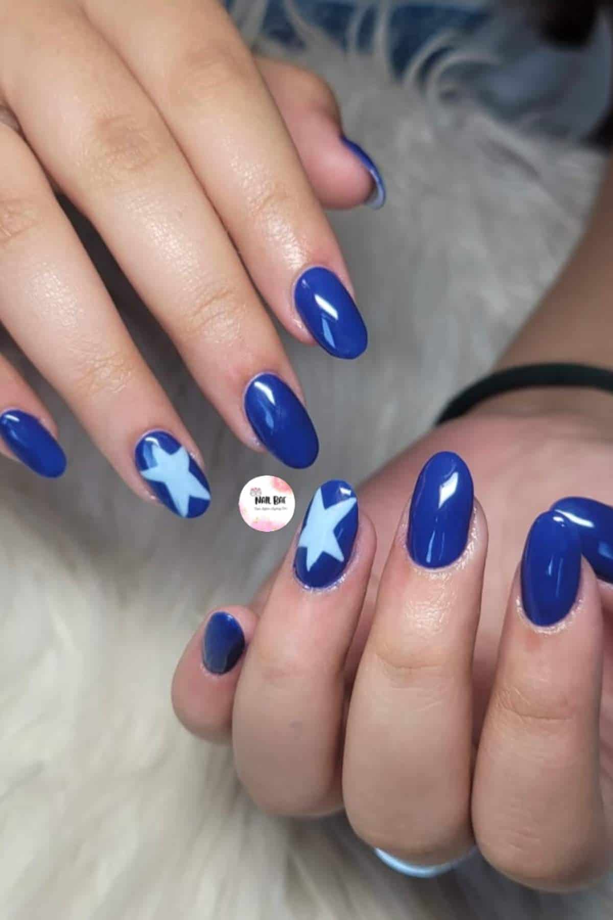 Hands with blue oval-shaped nails; two nails feature a white star design on a soft white surface background.