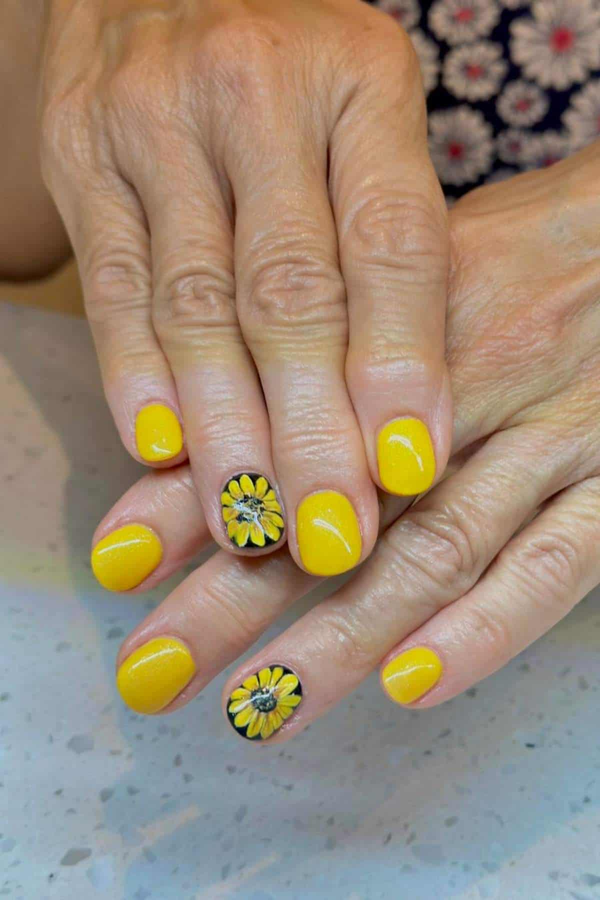 Hands with short, rounded nails painted yellow; the ring fingernails feature detailed yellow and black daisy flower designs.