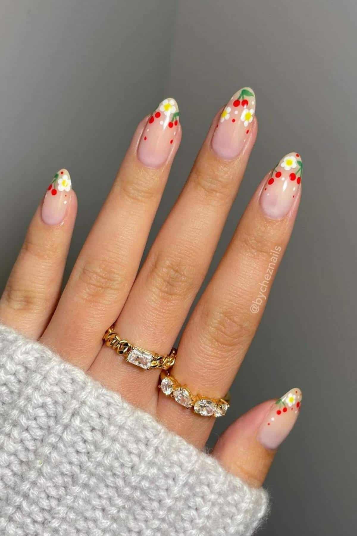 A hand with almond-shaped nails featuring a nude base, green tips, and red and white dot accents. The person wears two gold rings and a gray knit sweater.